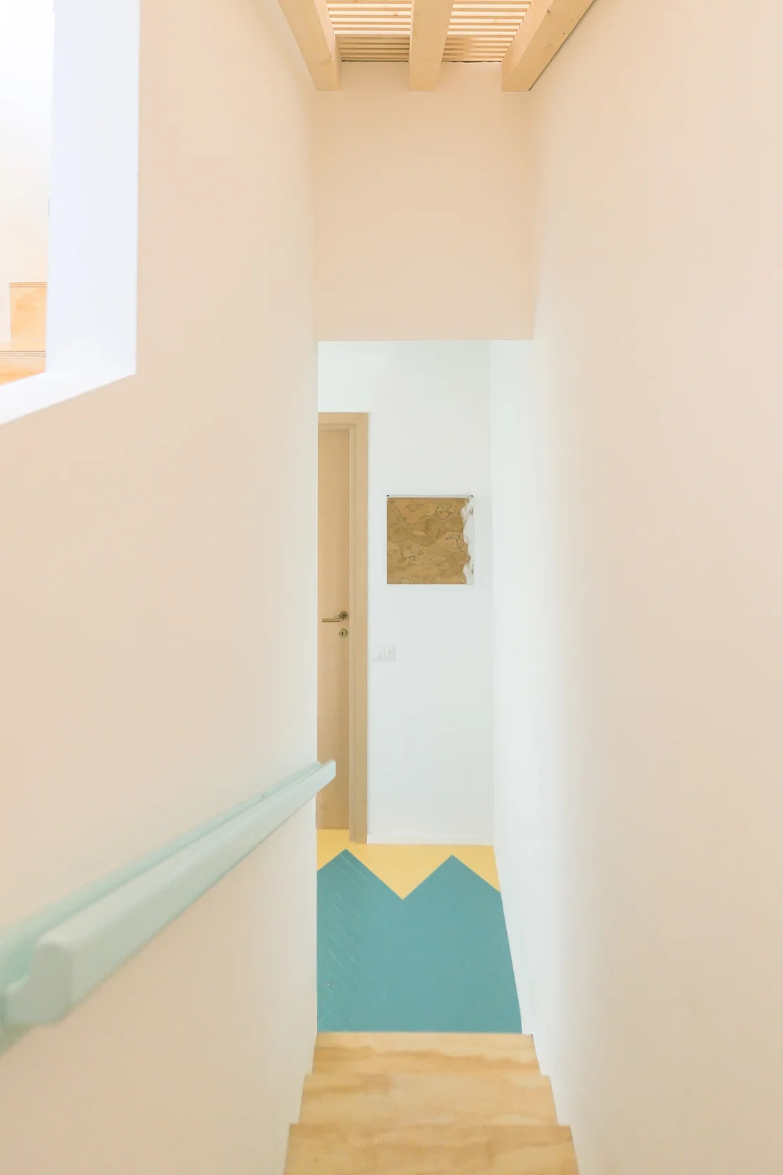 A white staircase with a light blue handrail leads down to a hallway featuring a bold yellow and teal chevron pattern floor.