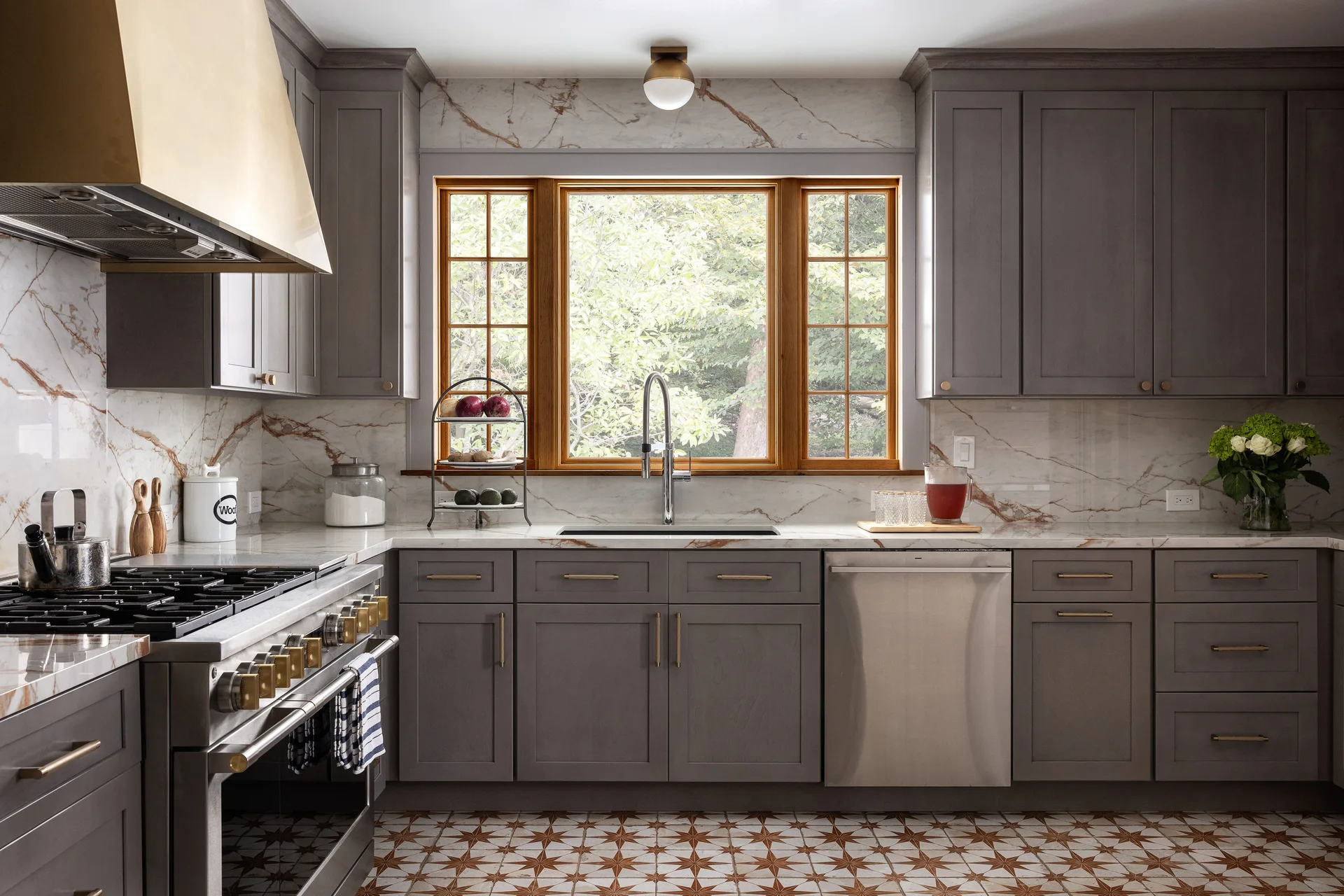 A kitchen with grey shaker cabinets, a marble slab backsplash, stainless steel appliances, a wood-framed window, and star-patterned floor tile.