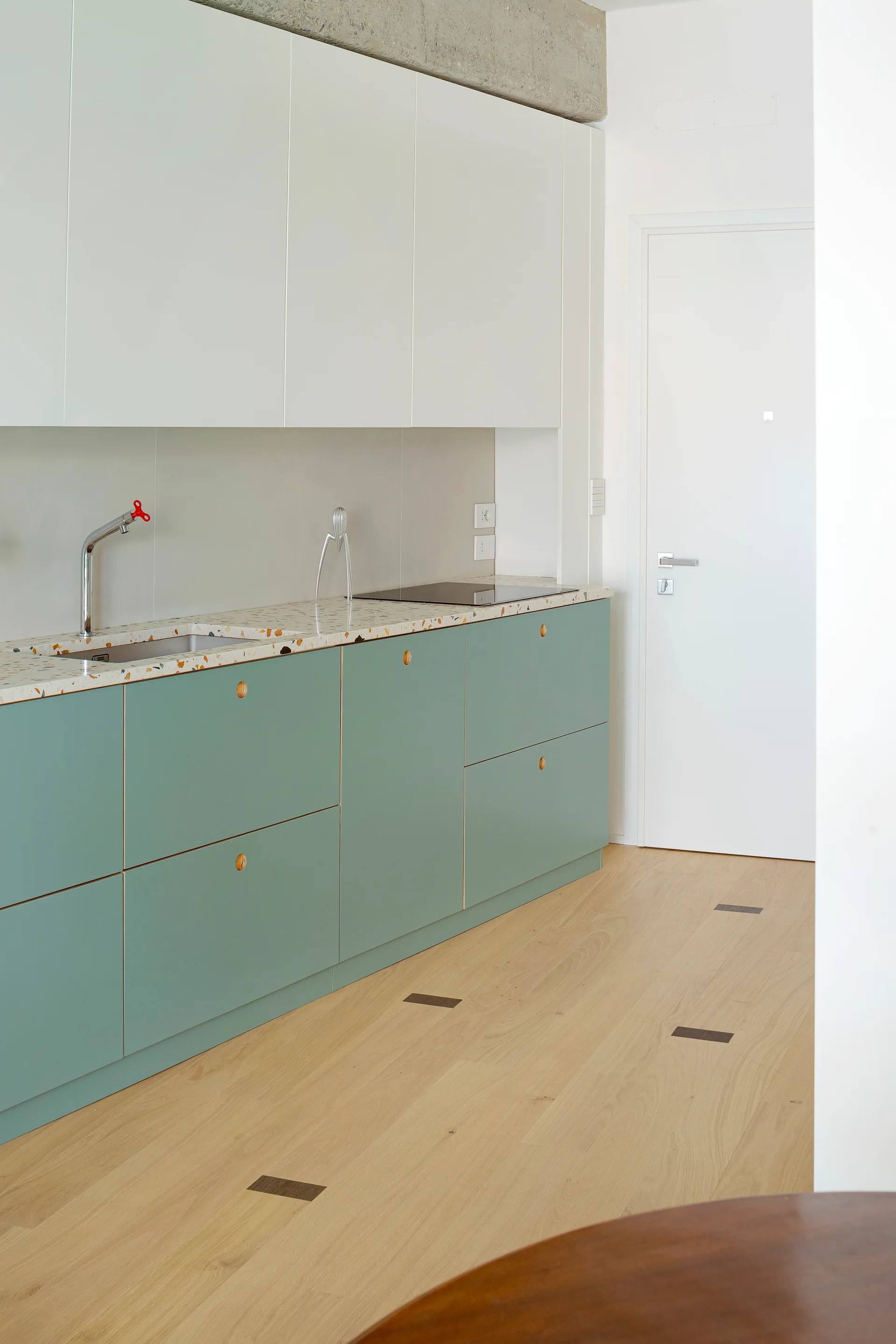 A modern kitchen with sage green lower cabinets, a speckled terrazzo countertop, white upper cabinetry, and light wood flooring.