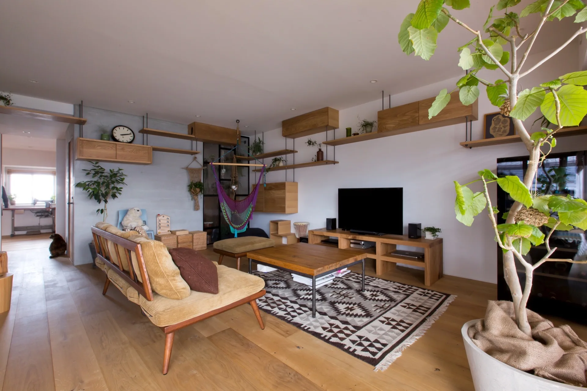 A residential living room features a tan corduroy sofa, a patterned rug under a wooden coffee table, and extensive wall-mounted wood shelves.