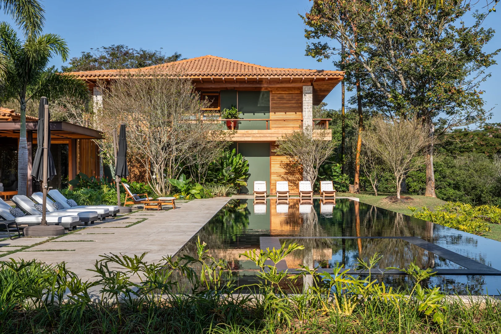 A two-story wooden house with a tiled roof stands behind a black-bottom swimming pool lined with white lounge chairs on a stone patio.