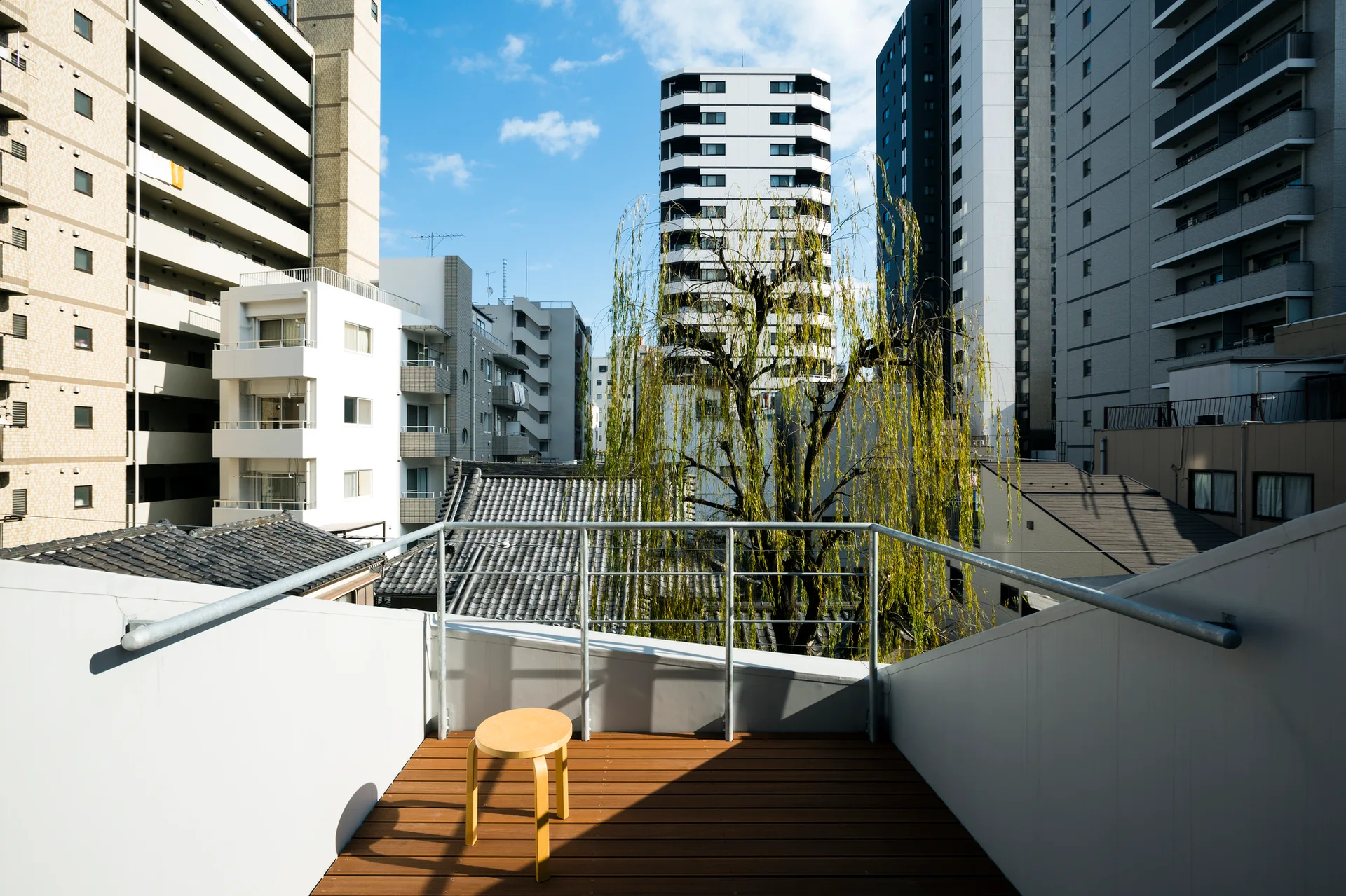 A simple wooden stool stands on a brown wood plank balcony deck with white walls and a metal railing overlooking city buildings.