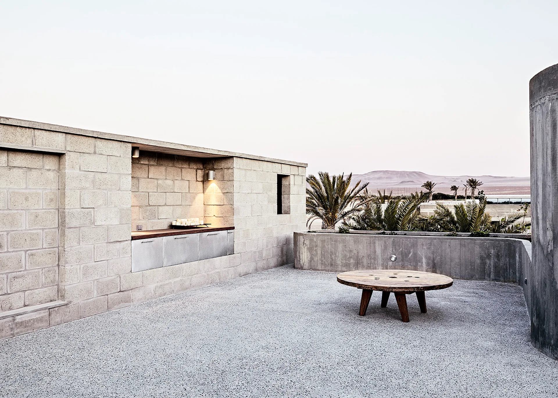 A minimalist concrete patio features a built-in kitchen niche with warm lighting, a low round wooden table, and a planter with palm trees.