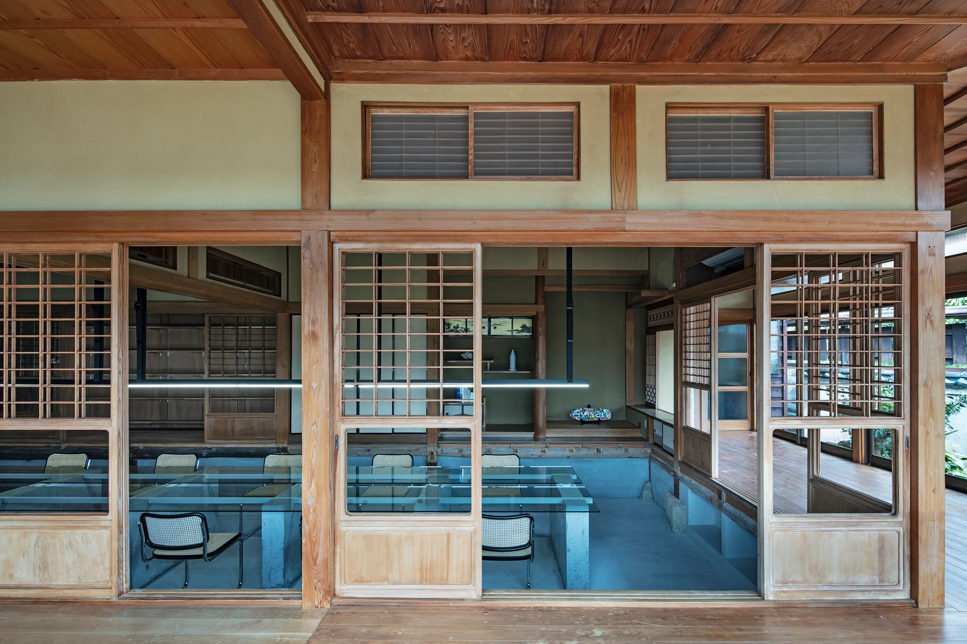Sliding wooden lattice doors frame a modern workspace featuring glass-topped tables, cane-back chairs, and concrete floors.