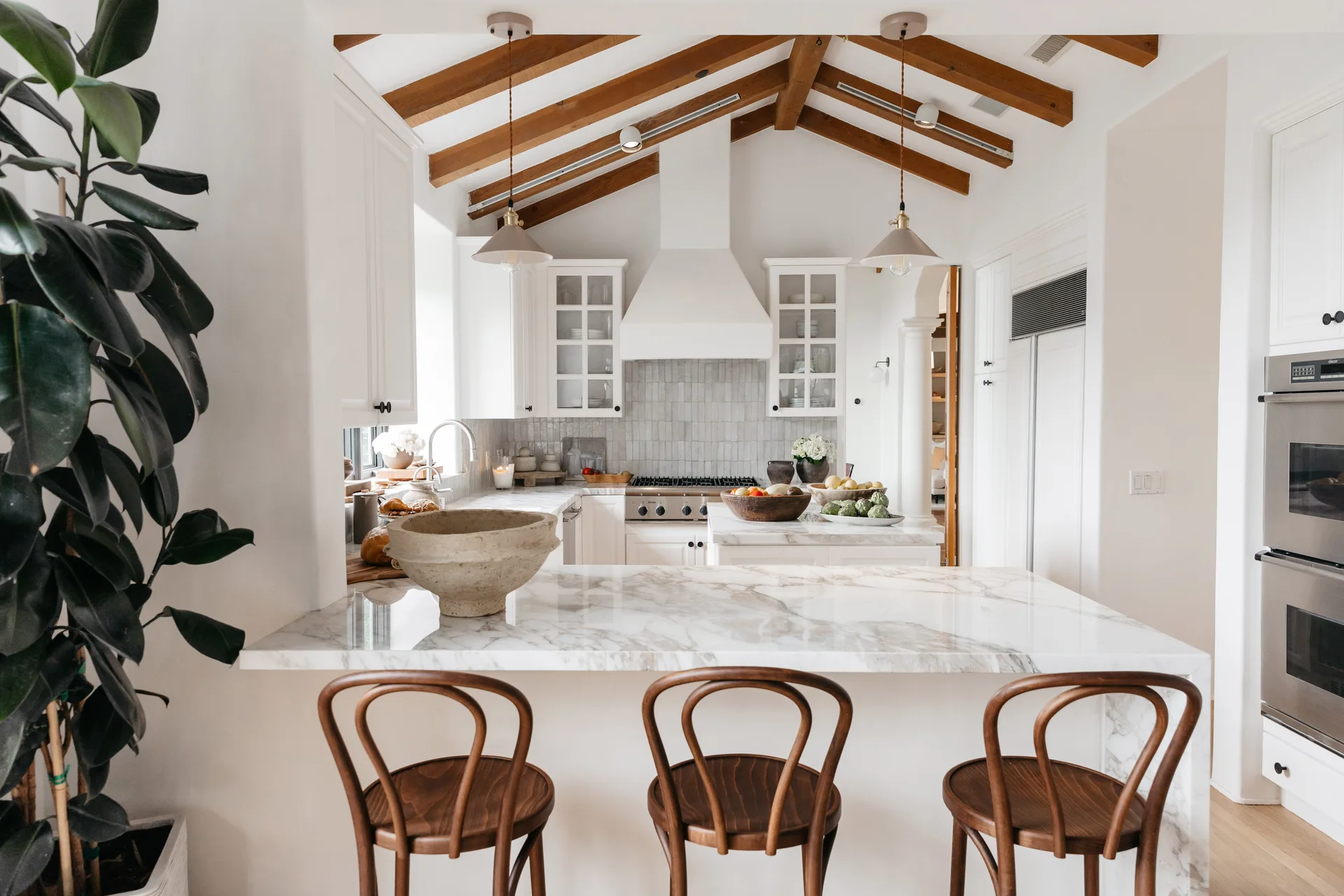 A bright kitchen features a white marble waterfall island with three wooden bentwood stools under high vaulted ceilings with exposed timber beams.