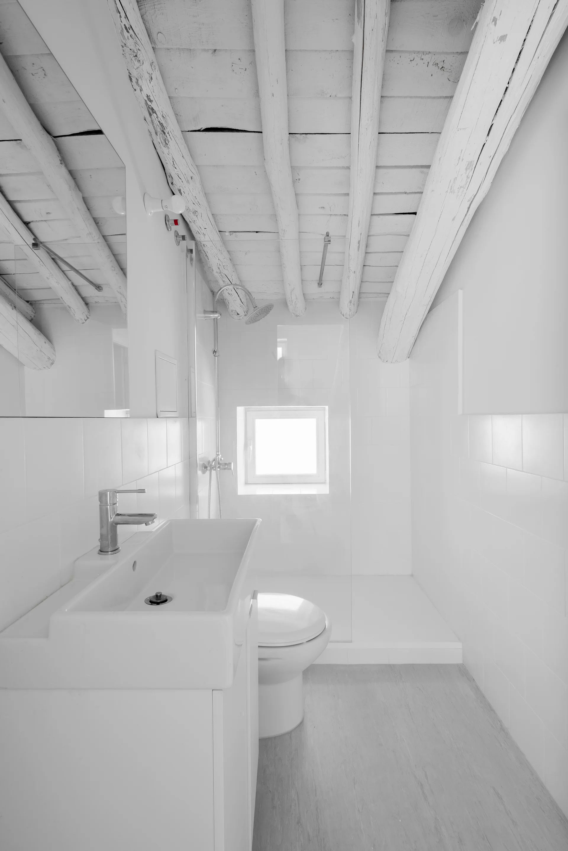 A compact white bathroom featuring exposed wooden ceiling beams, a rectangular ceramic sink, a toilet, and a walk-in shower with a glass partition.