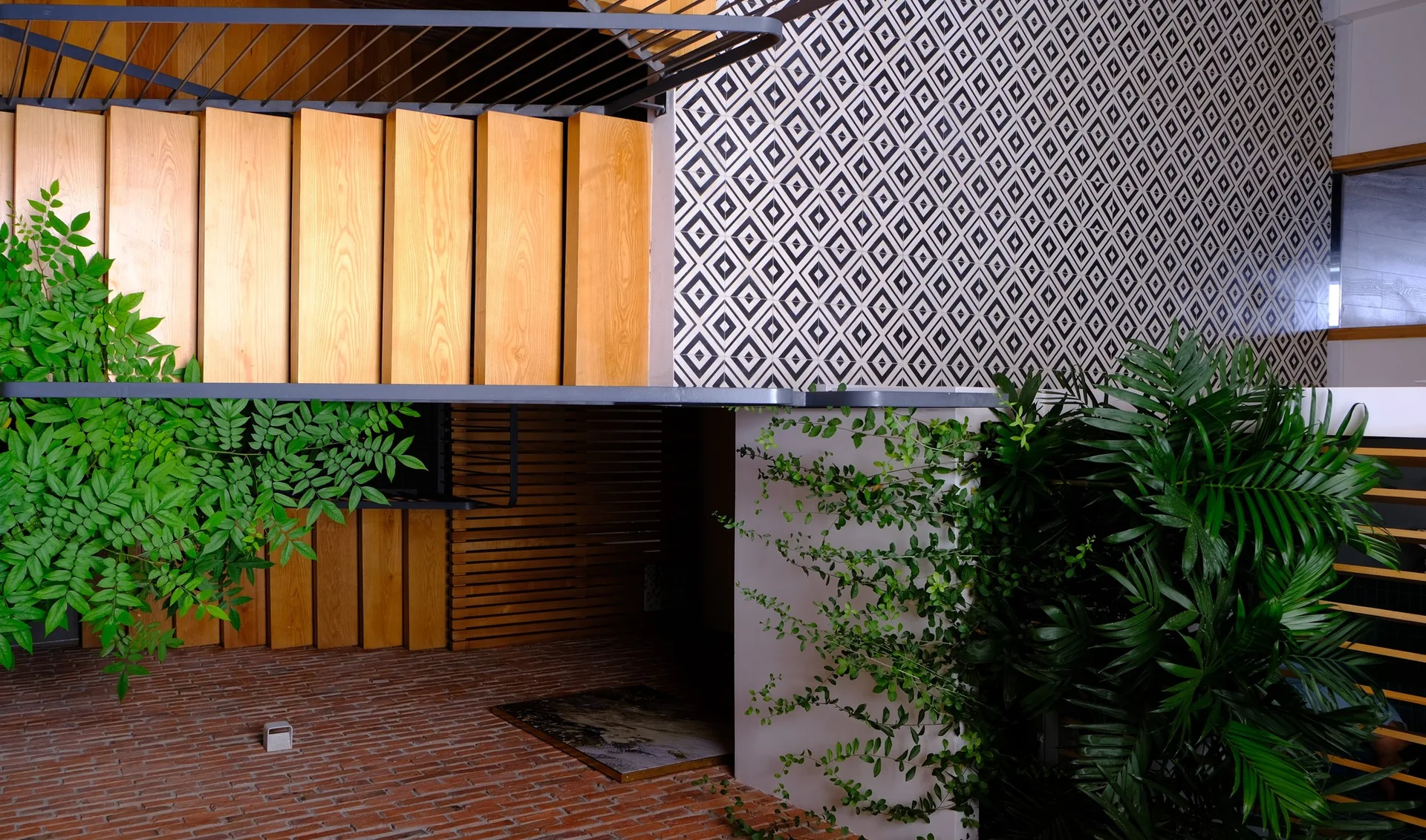 A modern indoor staircase with natural wood treads is flanked by a black and white geometric patterned wall and climbing green plants on a brick floor.