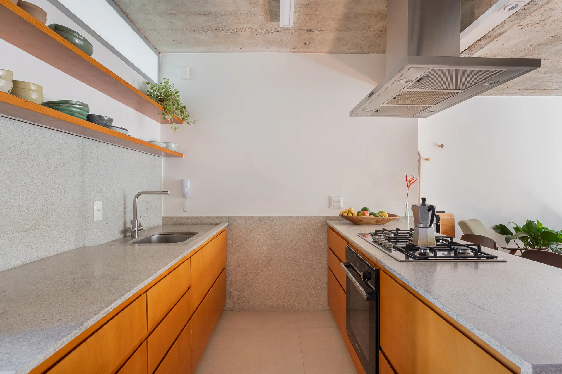 A modern galley kitchen features warm wood lower cabinets, light stone countertops, a stainless steel range hood, and an exposed concrete ceiling.
