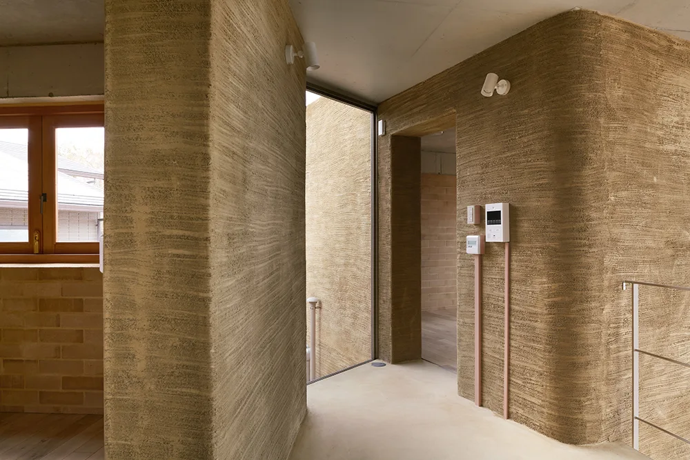 A minimalist hallway features heavily textured tan plaster walls with curved corners beneath a raw concrete ceiling with white spotlight fixtures.