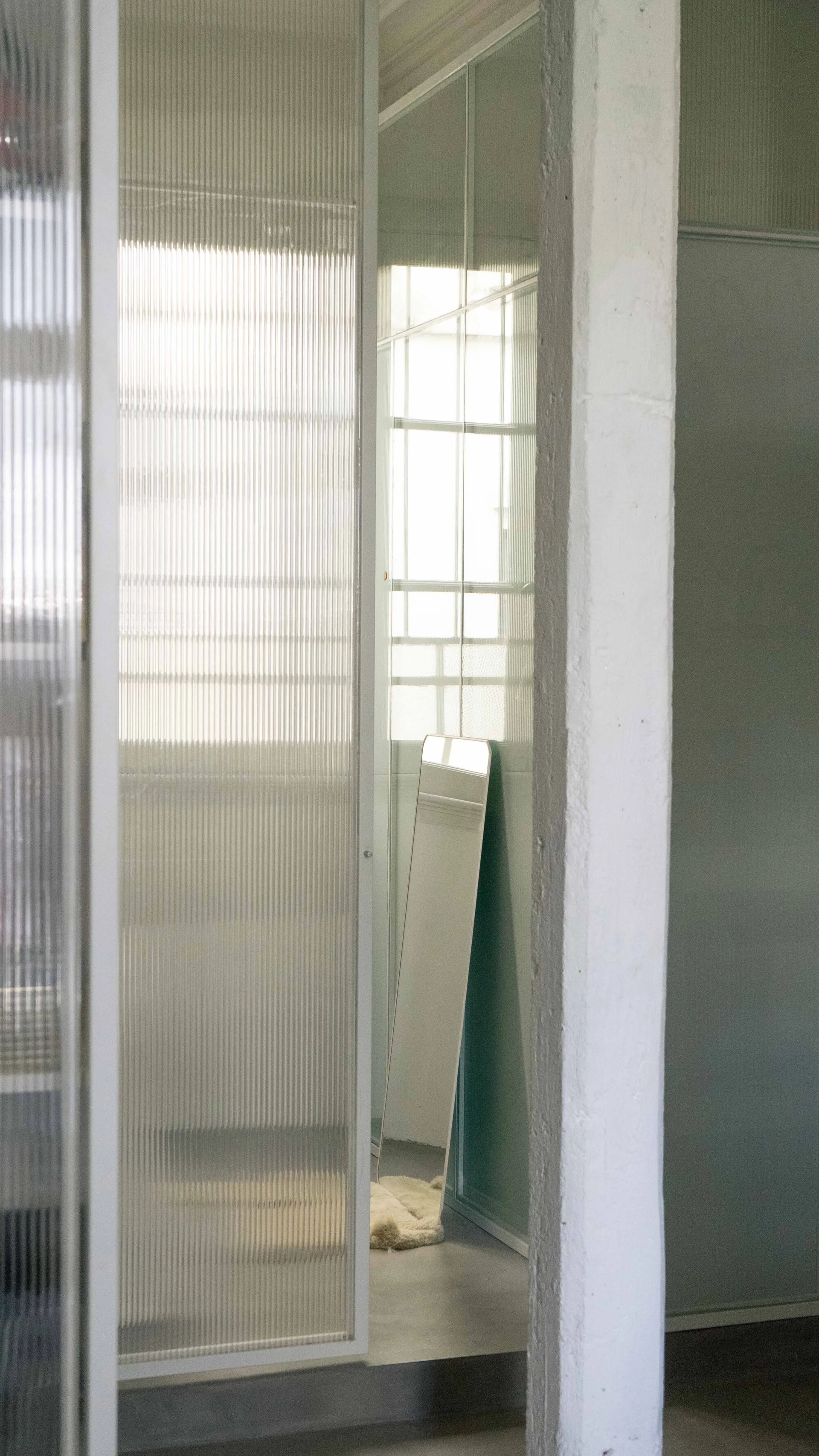 A white structural column stands beside sliding room divider panels made of ribbed fluted glass, with a slim mirror leaning in the background.