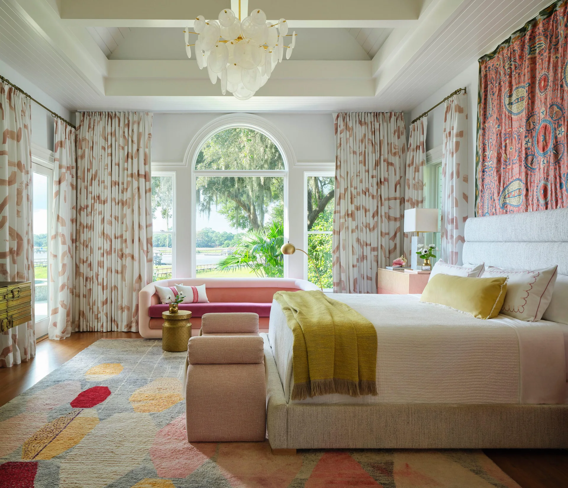 A bright bedroom featuring an upholstered bed, a curved pink sofa by a large arched window, patterned drapes, and a colorful geometric rug.