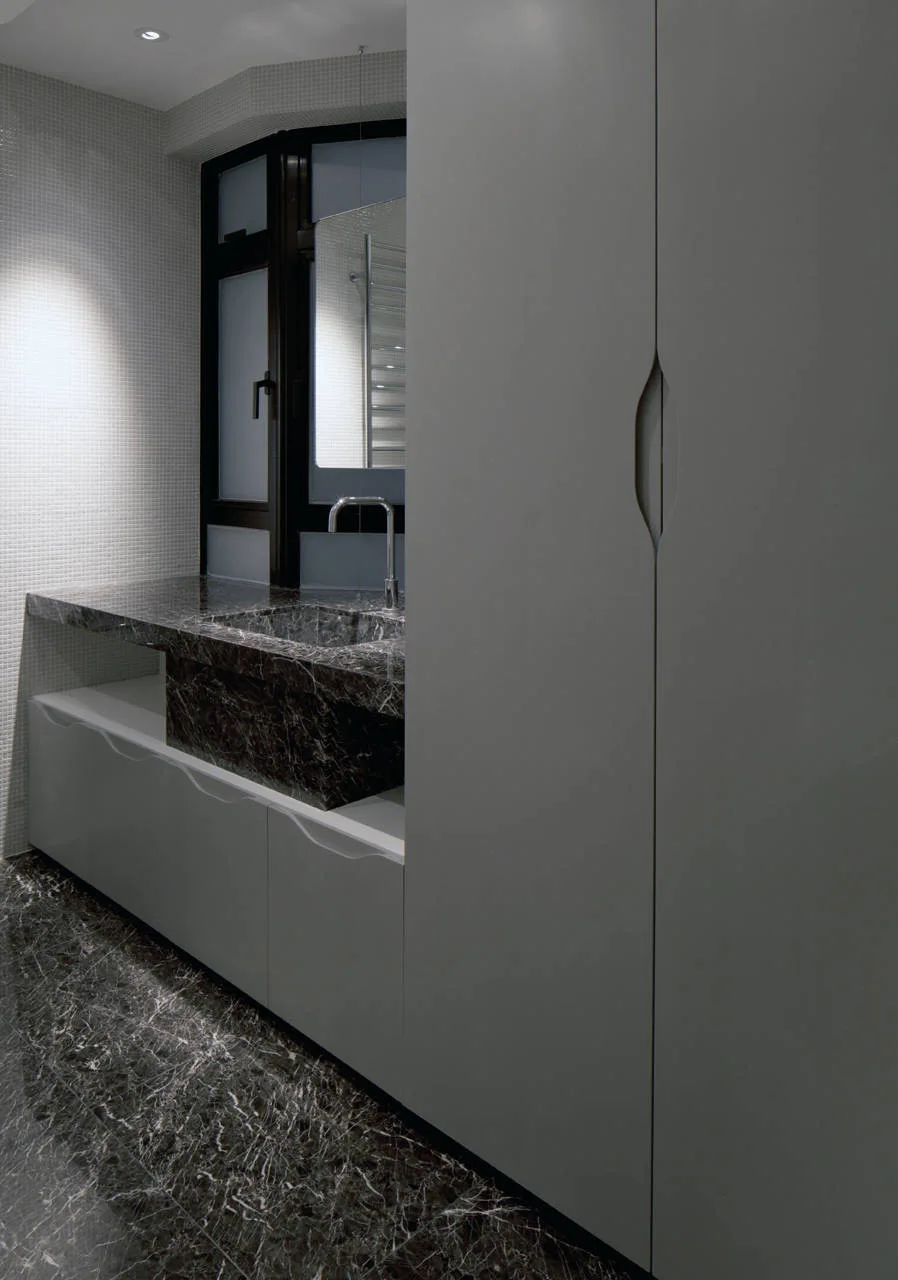 A modern bathroom features a dark marble vanity and floor, white mosaic tile walls, and sleek grey cabinetry with a sculptural cutout handle.