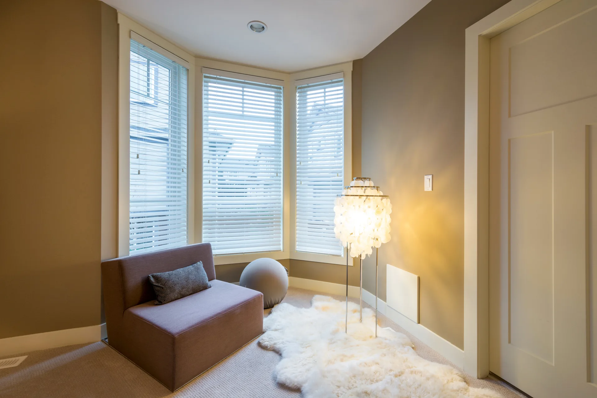 A small reading nook with a brown armless chair, a glowing shell floor lamp, and a white faux fur rug in a bay window.