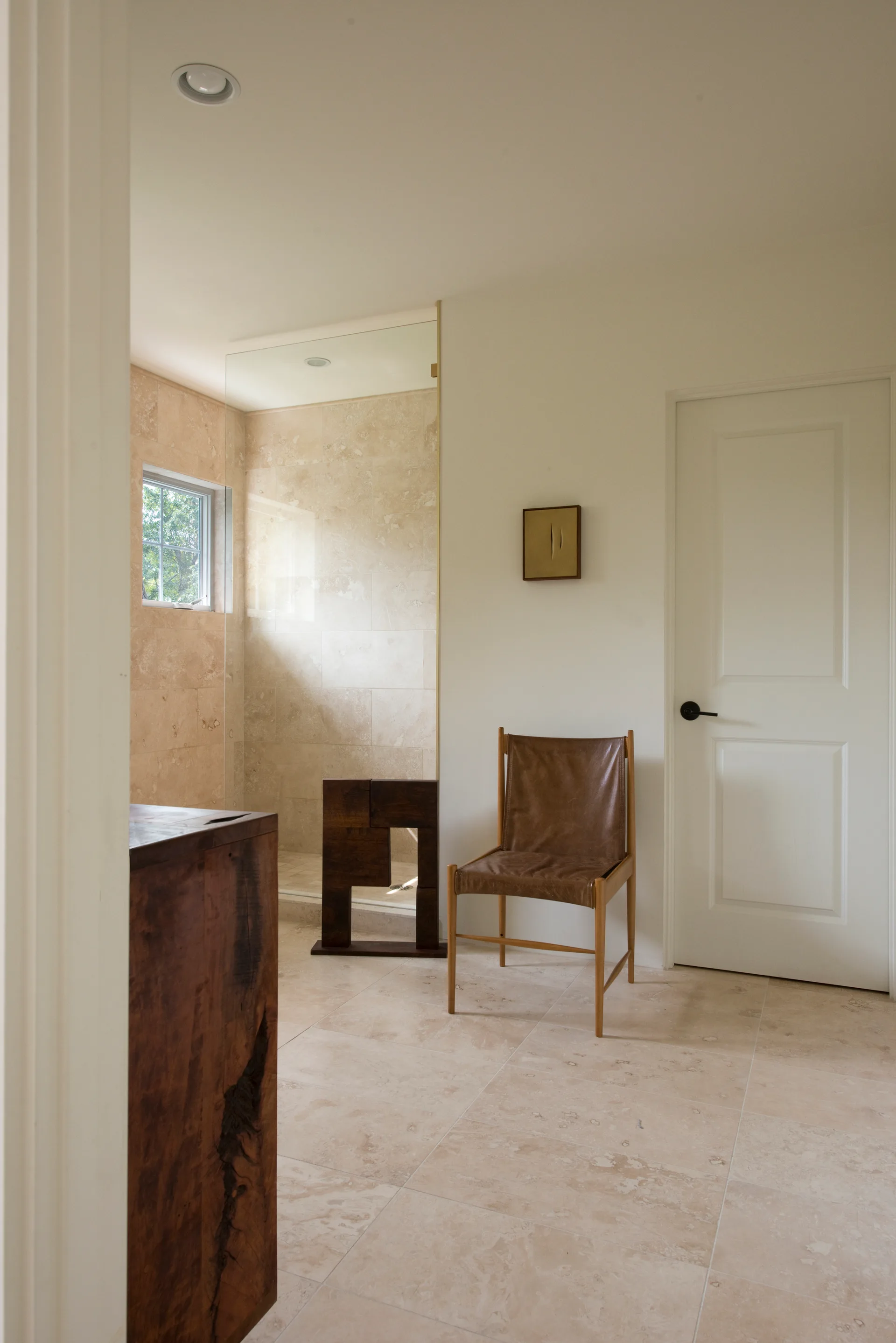 A minimalist bathroom features a glass-enclosed stone shower, a brown leather sling chair, and large beige floor tiles.
