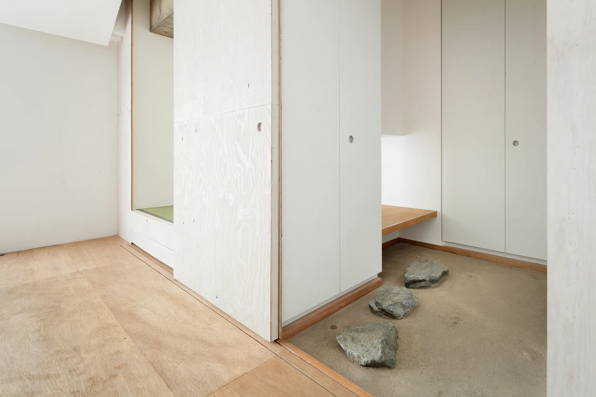 A minimalist entryway featuring white washed plywood wall panels, a wood plank floor, and stepping stones laid over a concrete transition zone.