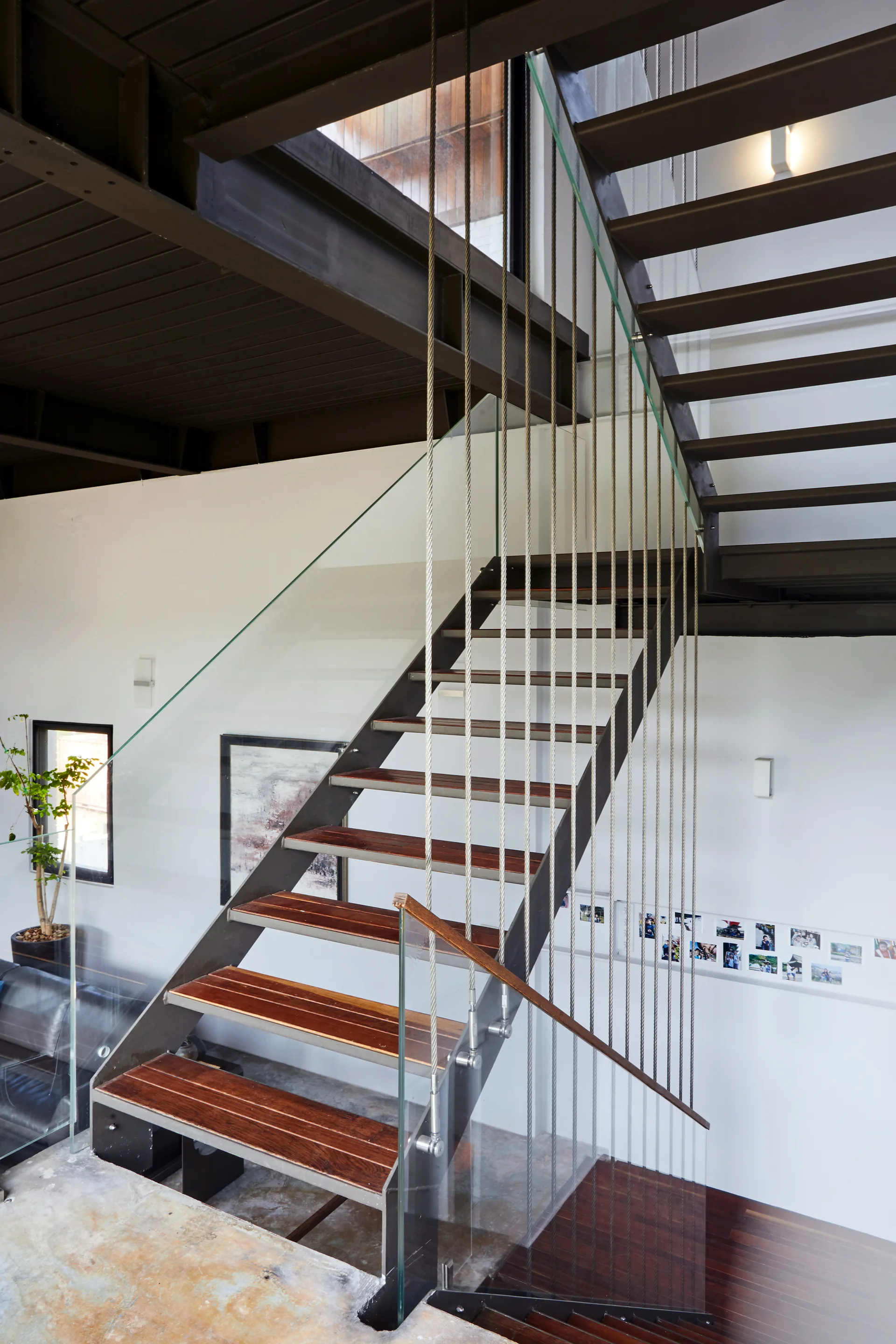A modern industrial staircase features dark wood treads, a structural steel frame, vertical metal cables, and glass panel safety barriers.