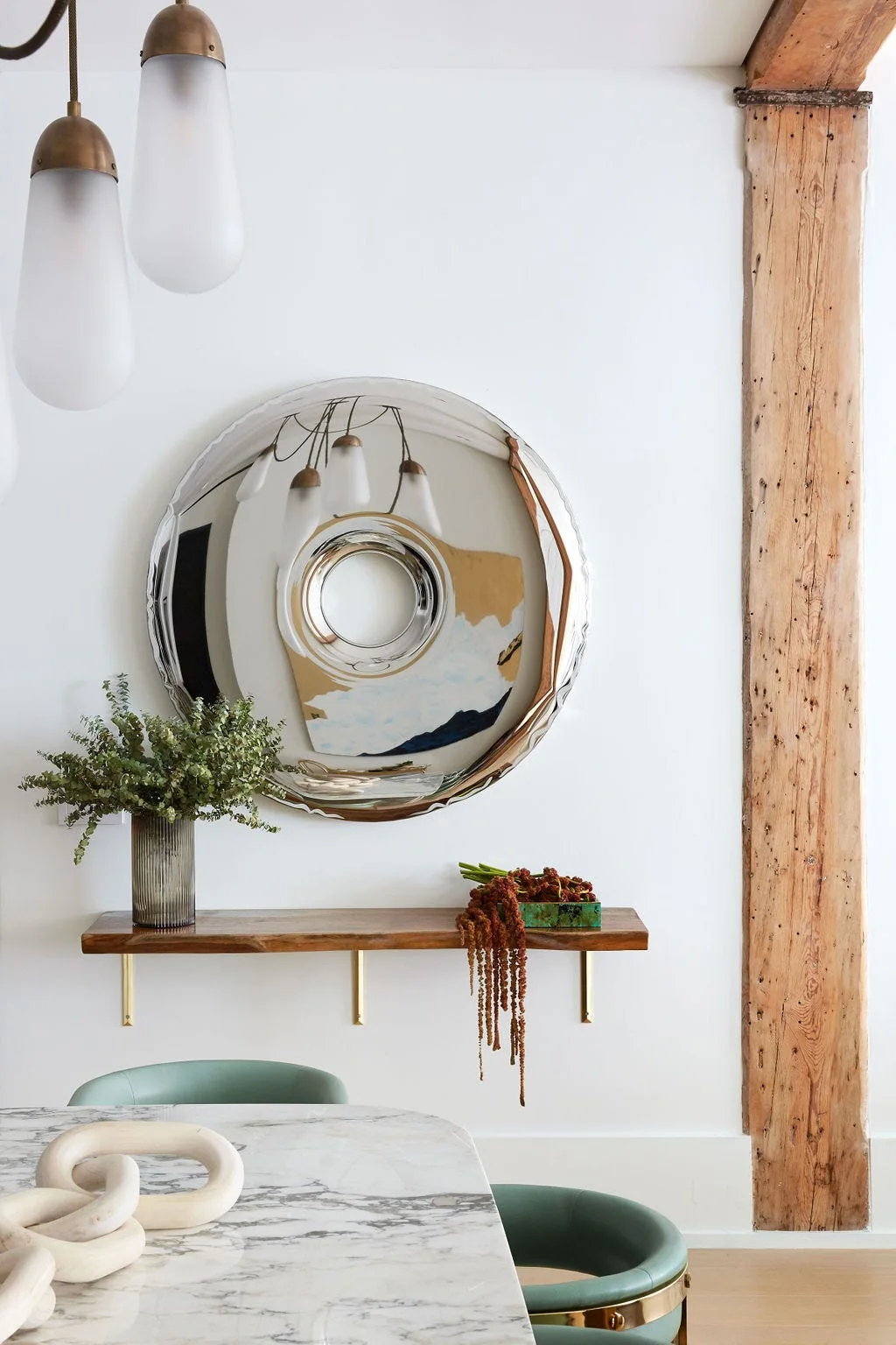 A round metallic convex mirror hangs above a floating wooden shelf on a white wall next to a rustic wooden column and a marble table.