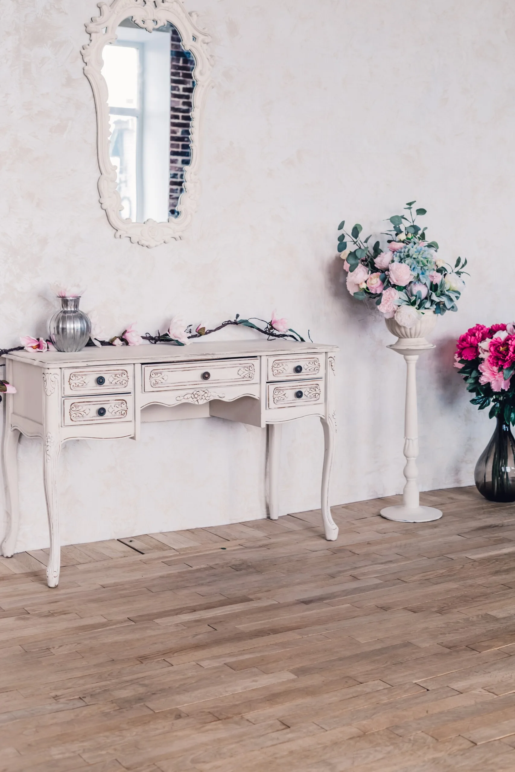 An ornate white vintage console table sits against a textured wall under a carved mirror, flanked by colorful floral arrangements on wood flooring.