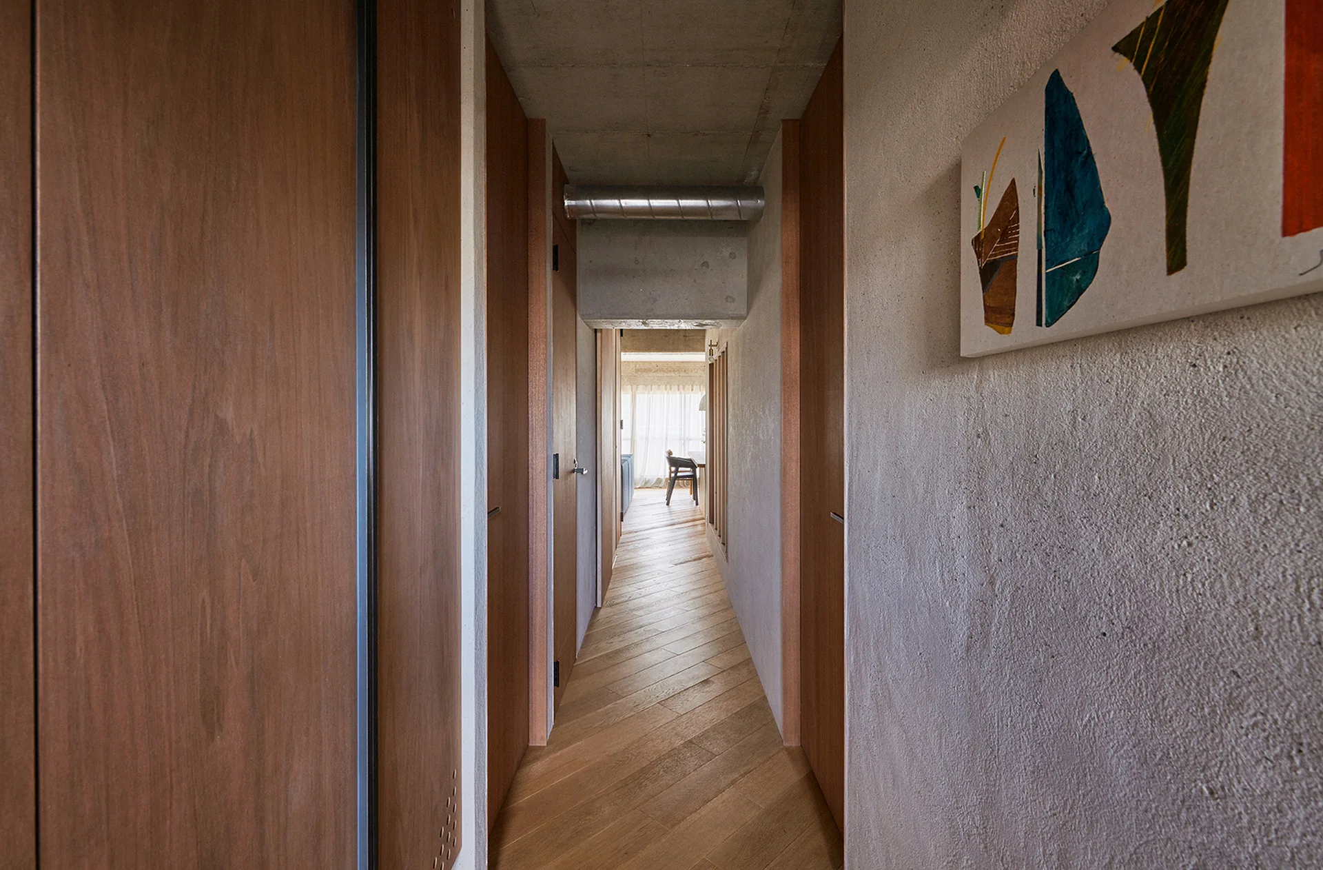 A narrow modern hallway featuring chevron wood flooring, warm wood-paneled doors, textured plaster walls, and a raw concrete ceiling.