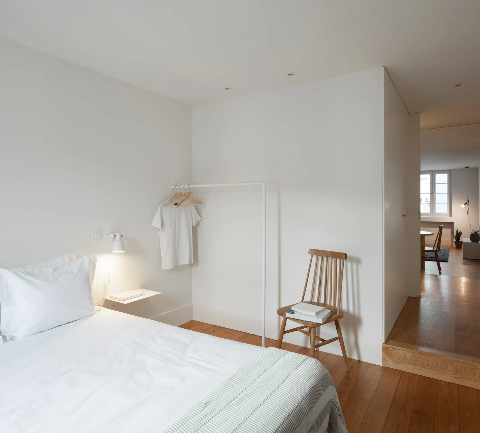 A minimalist white bedroom features hardwood flooring, a wooden spindle chair, a white metal clothing rack, and a bed with a warm wall sconce.