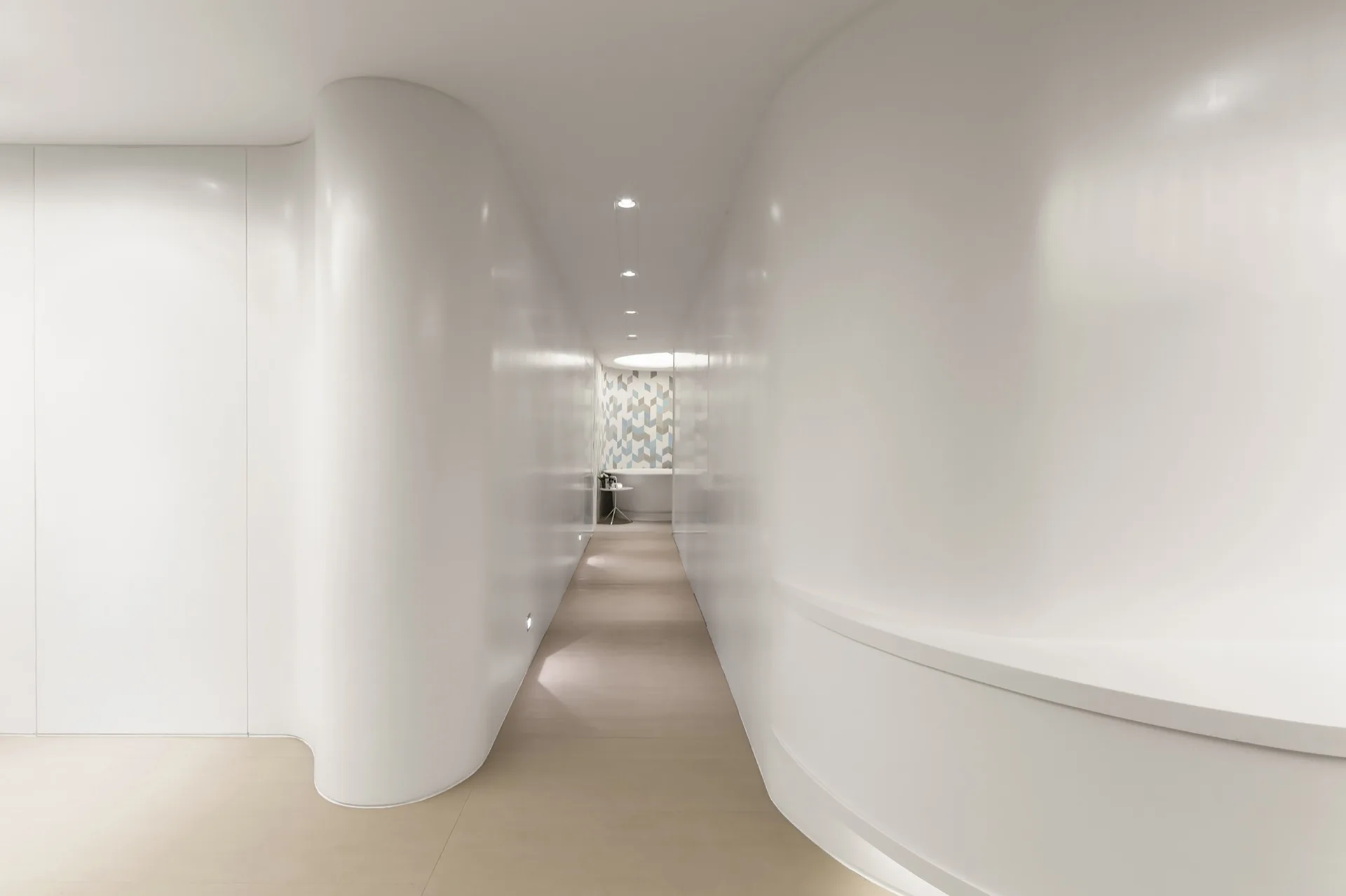 A long minimalist hallway features high-gloss curved white walls, recessed ceiling spotlights, and a geometric tiled feature wall at the far end.