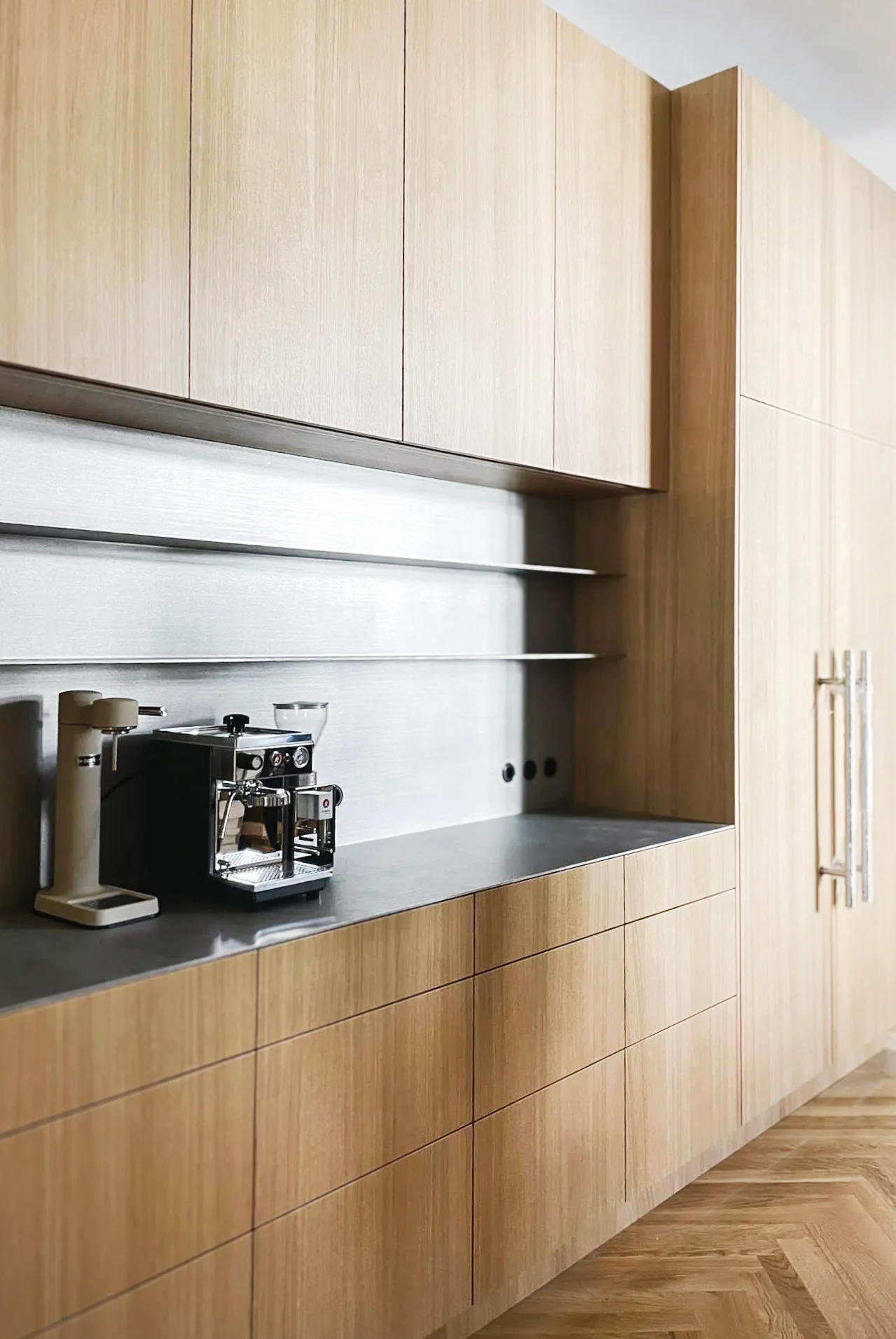 Light wood kitchen cabinets frame a dark matte countertop with an espresso machine against a brushed metal backsplash and herringbone floor.