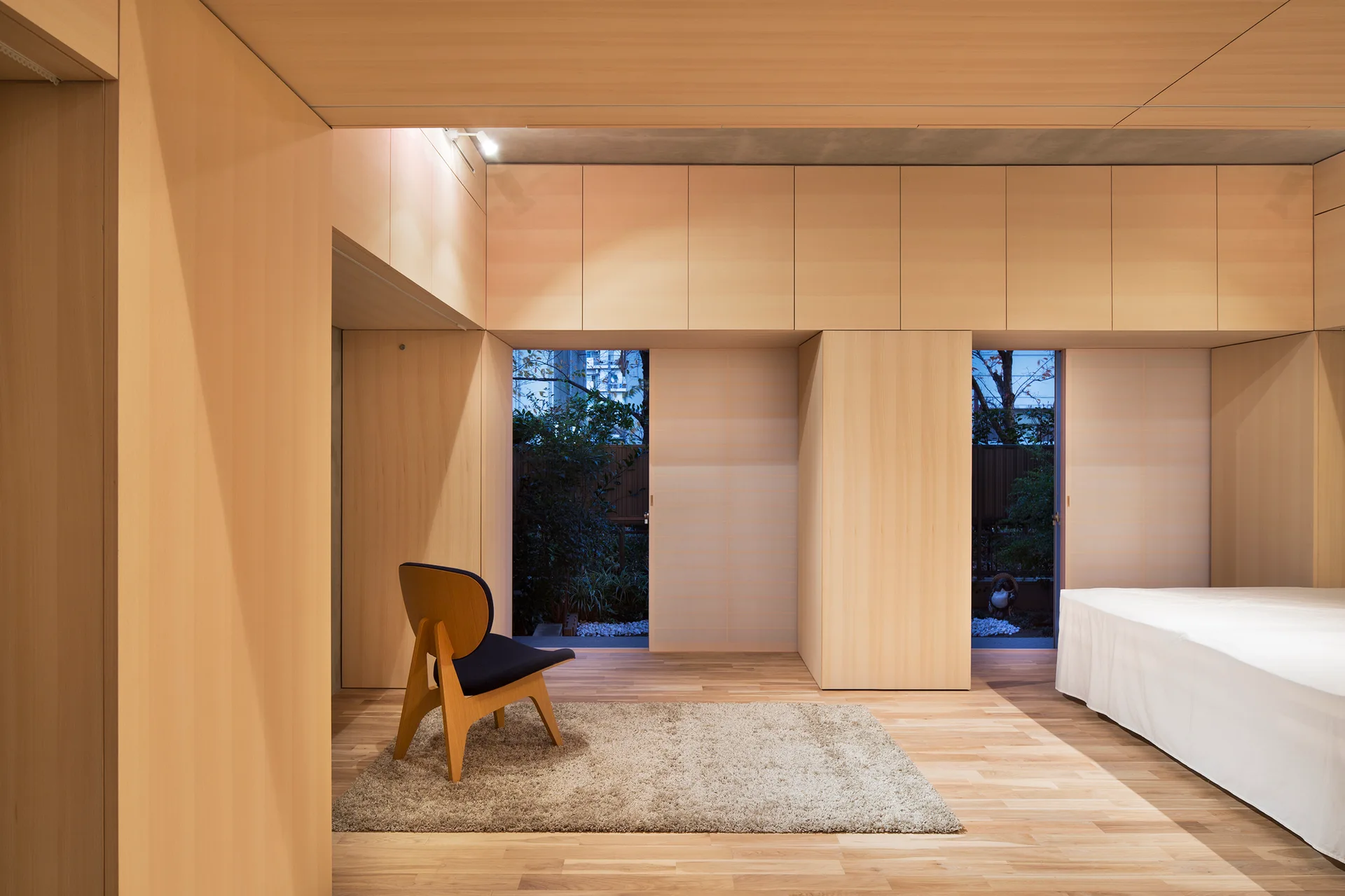 Minimalist bedroom with floor-to-ceiling light wood cabinetry, a wooden accent chair on a small rug, and sliding doors opening to the garden.