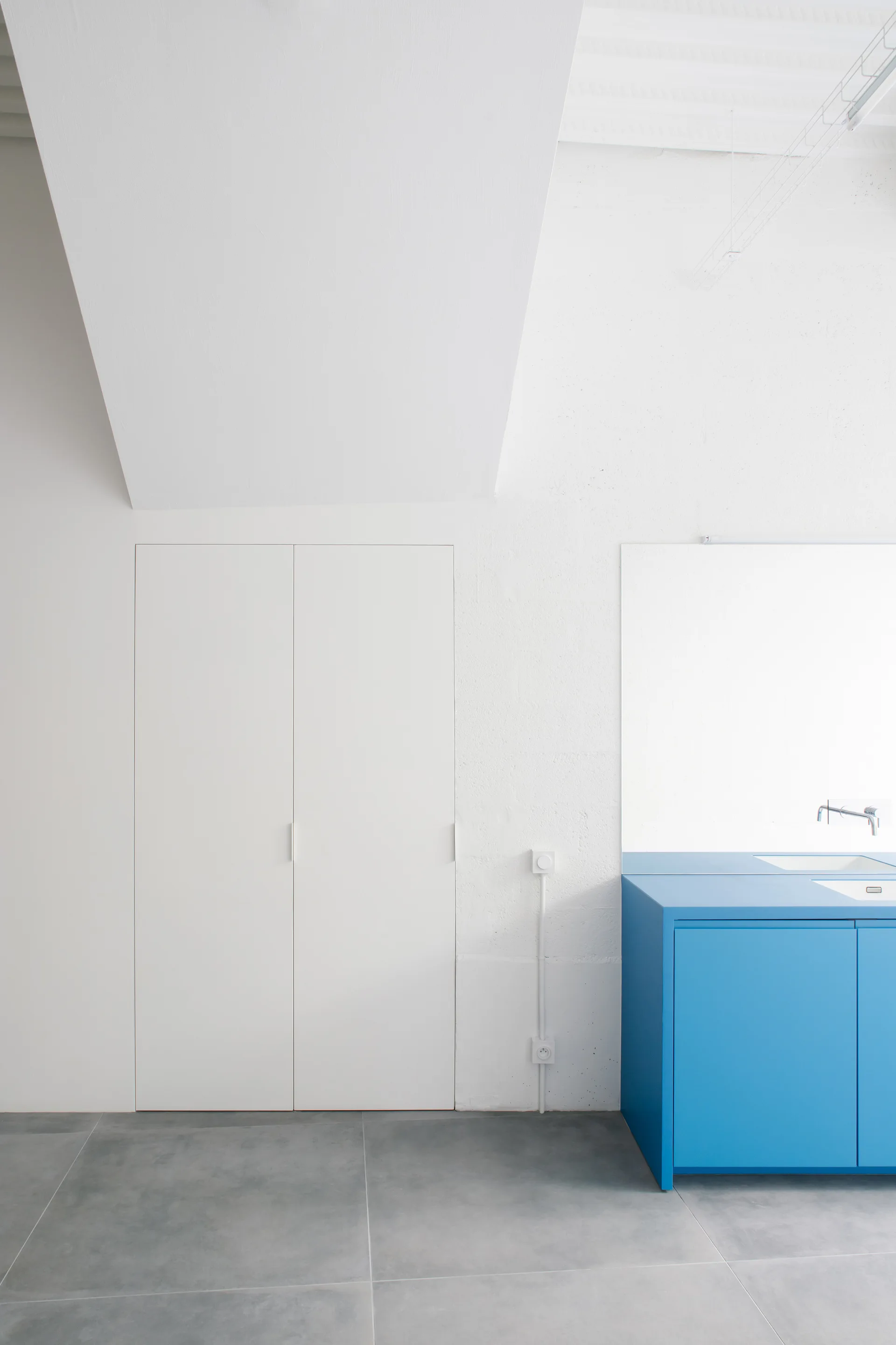 A minimalist white kitchen features a vibrant blue cabinet with an integrated sink, flush white closet doors, and large gray floor tiles.