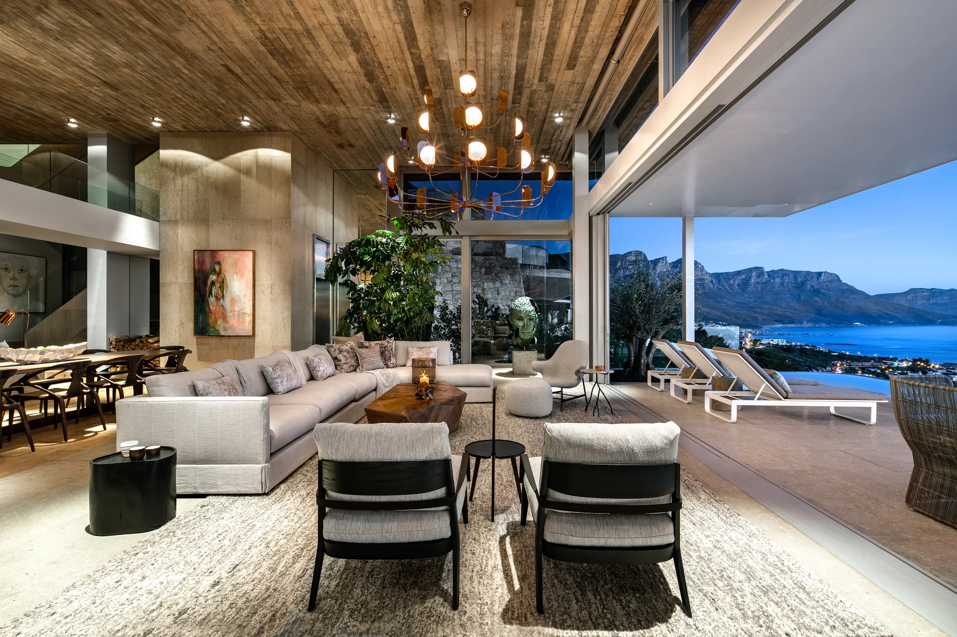 A modern living room features a large gray sectional sofa, a wood-paneled ceiling, and sliding glass doors opening to an outdoor terrace overlooking the sea.