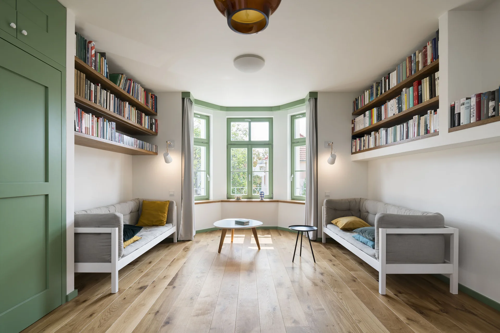 Two gray sofas face each other under wooden bookshelves in a white living room featuring a green-trimmed bay window and light hardwood floors.