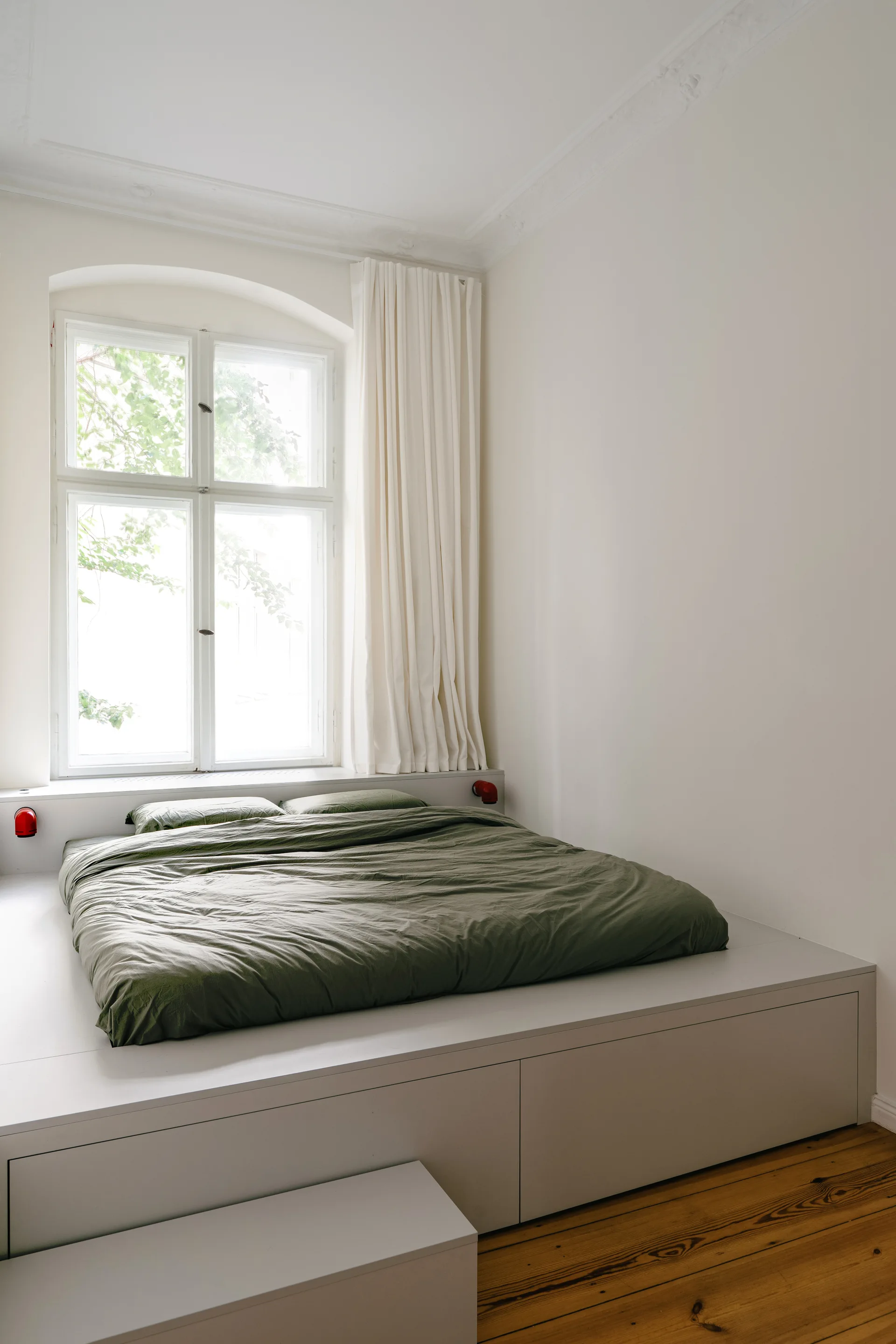 An olive green duvet rests on a white built-in platform bed with storage drawers below an arched window with white curtains.