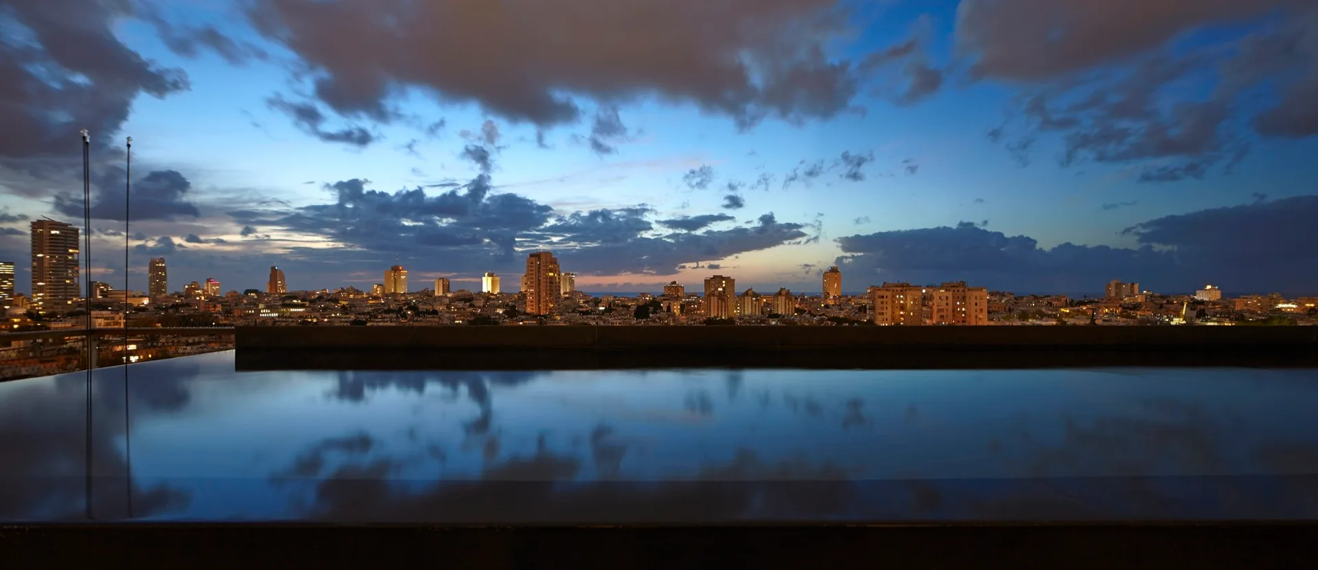 An infinity edge swimming pool reflects the dark blue sky and distant glowing city lights at twilight.