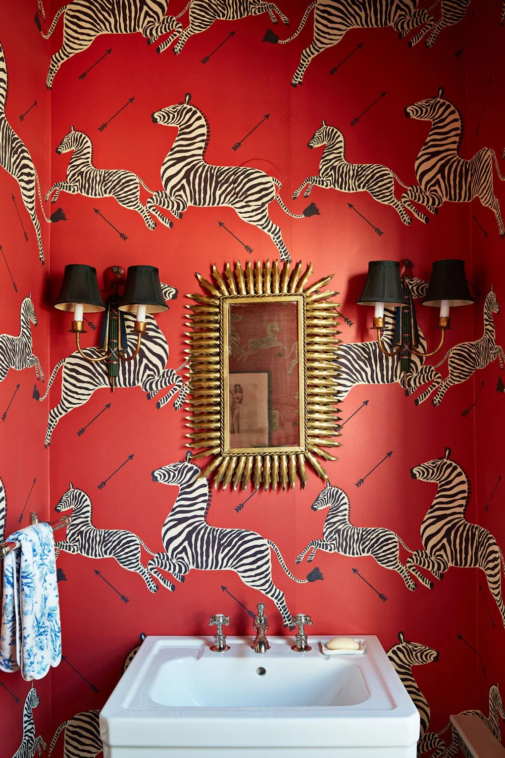 A powder room features bold red wallpaper with a zebra pattern, a gold sunburst mirror, and two black sconces above a white sink.