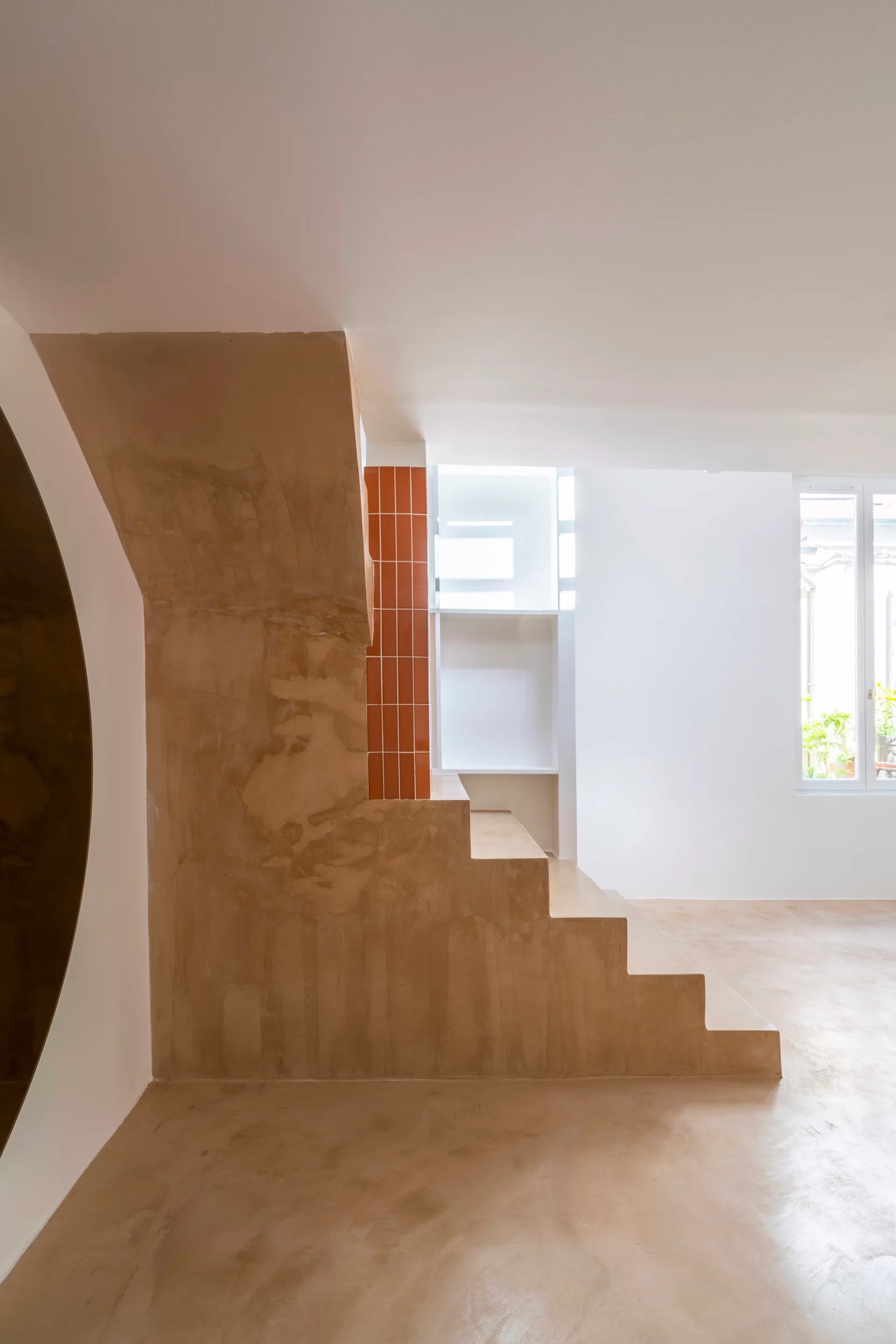 A minimalist tan plaster staircase stands next to a vertical strip of orange terracotta tiles in a white room with recessed shelving.