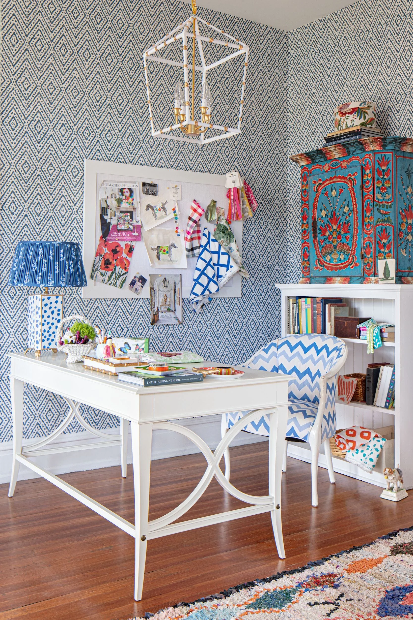 A home office features a white wooden desk, a blue and white chevron armchair, geometric wallpaper, and a lantern pendant light.
