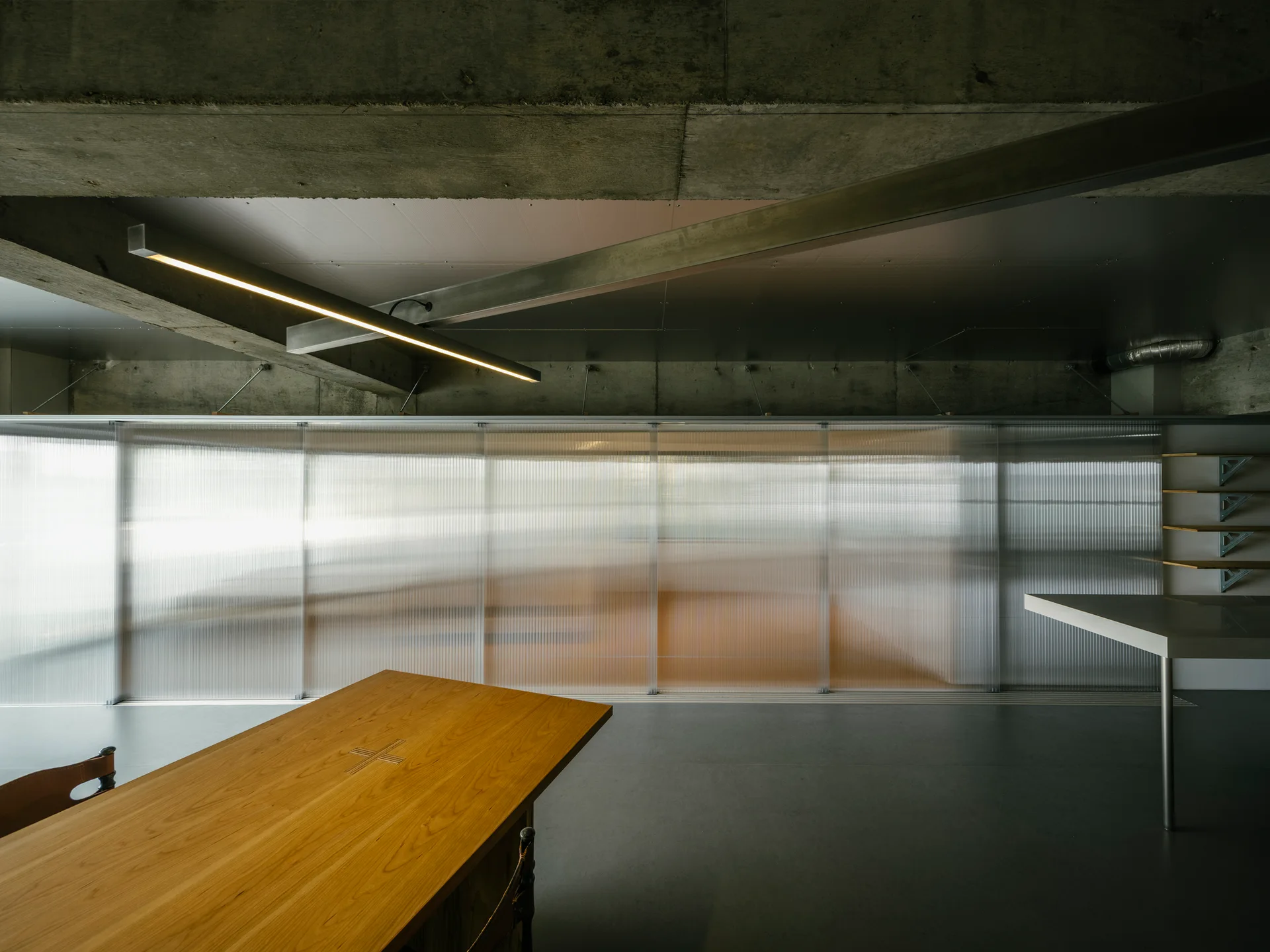 A wooden dining table sits in an industrial room featuring a raw concrete ceiling, a linear light fixture, and translucent ribbed sliding wall panels.