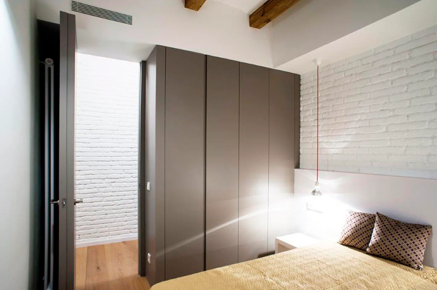 A modern bedroom features a large grey wardrobe, a white painted brick wall, and exposed wood beams on the ceiling.