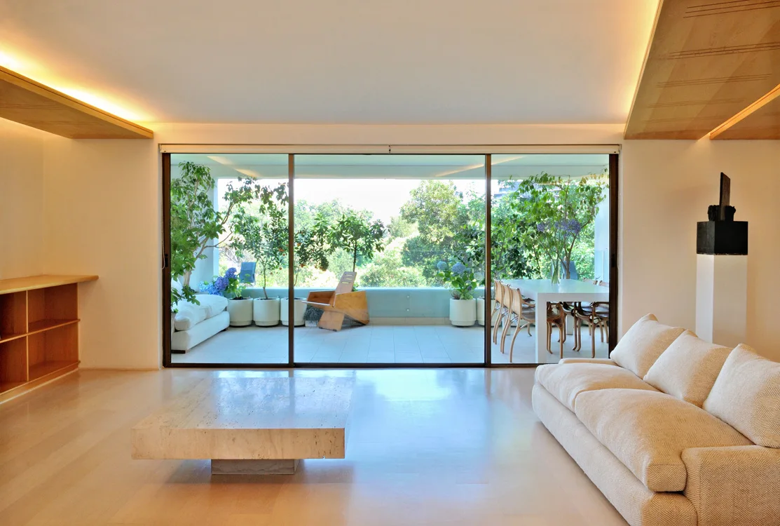A minimalist living room features a low travertine coffee table, a cream sofa, and sliding glass doors that open to a green terrace.