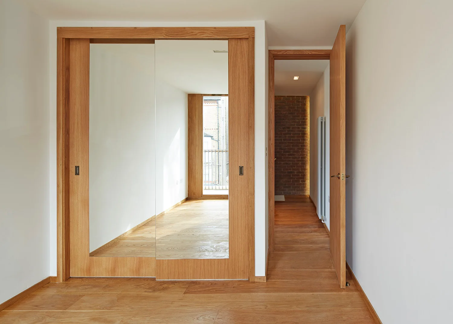 A minimalist bedroom featuring warm wood flooring, white walls, and a large wood-framed sliding mirrored closet next to an open doorway.