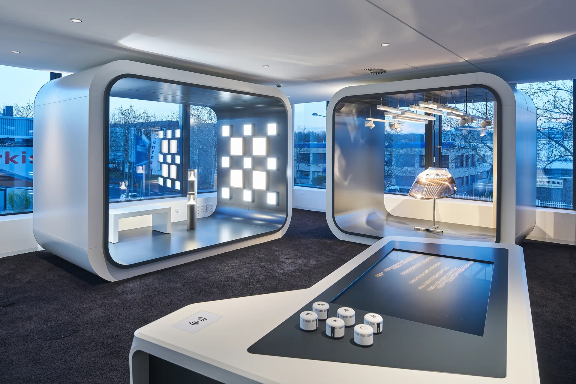 A modern showroom featuring two large curved modular display pods, an interactive touch screen table, and large background windows.