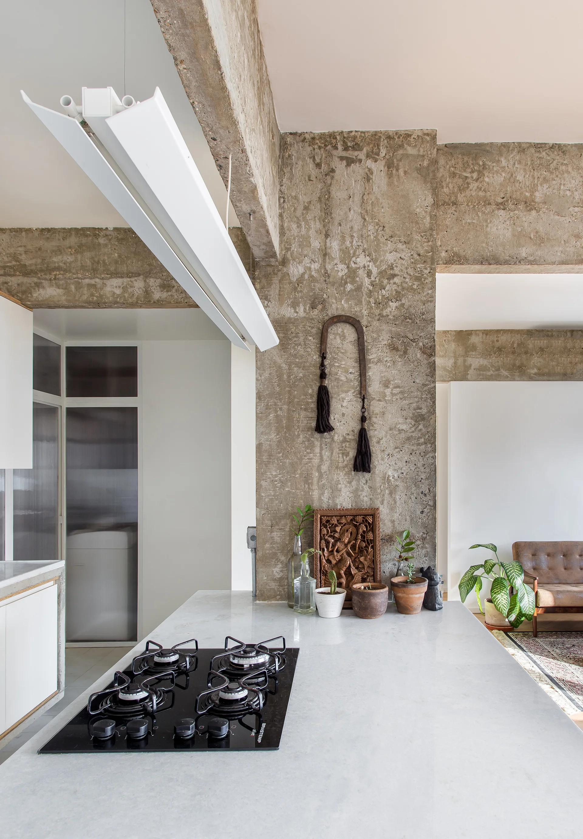 A kitchen island with a black gas cooktop is positioned next to a tall, textured concrete pillar decorated with small plants and a wood carving.