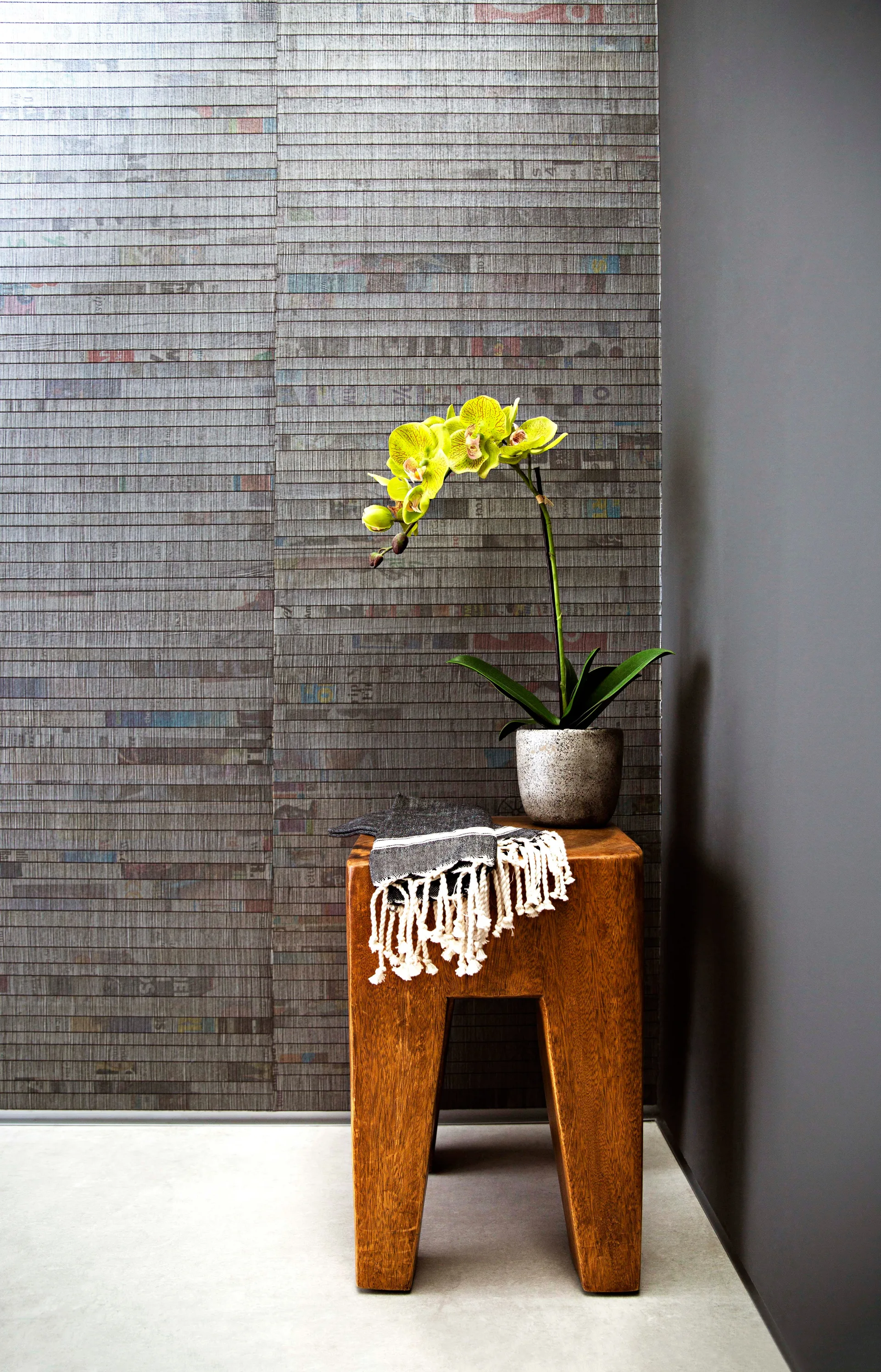 A rustic wooden stool holds a potted yellow orchid and a fringed black textile against a textured grey newspaper-print wallpapered wall.