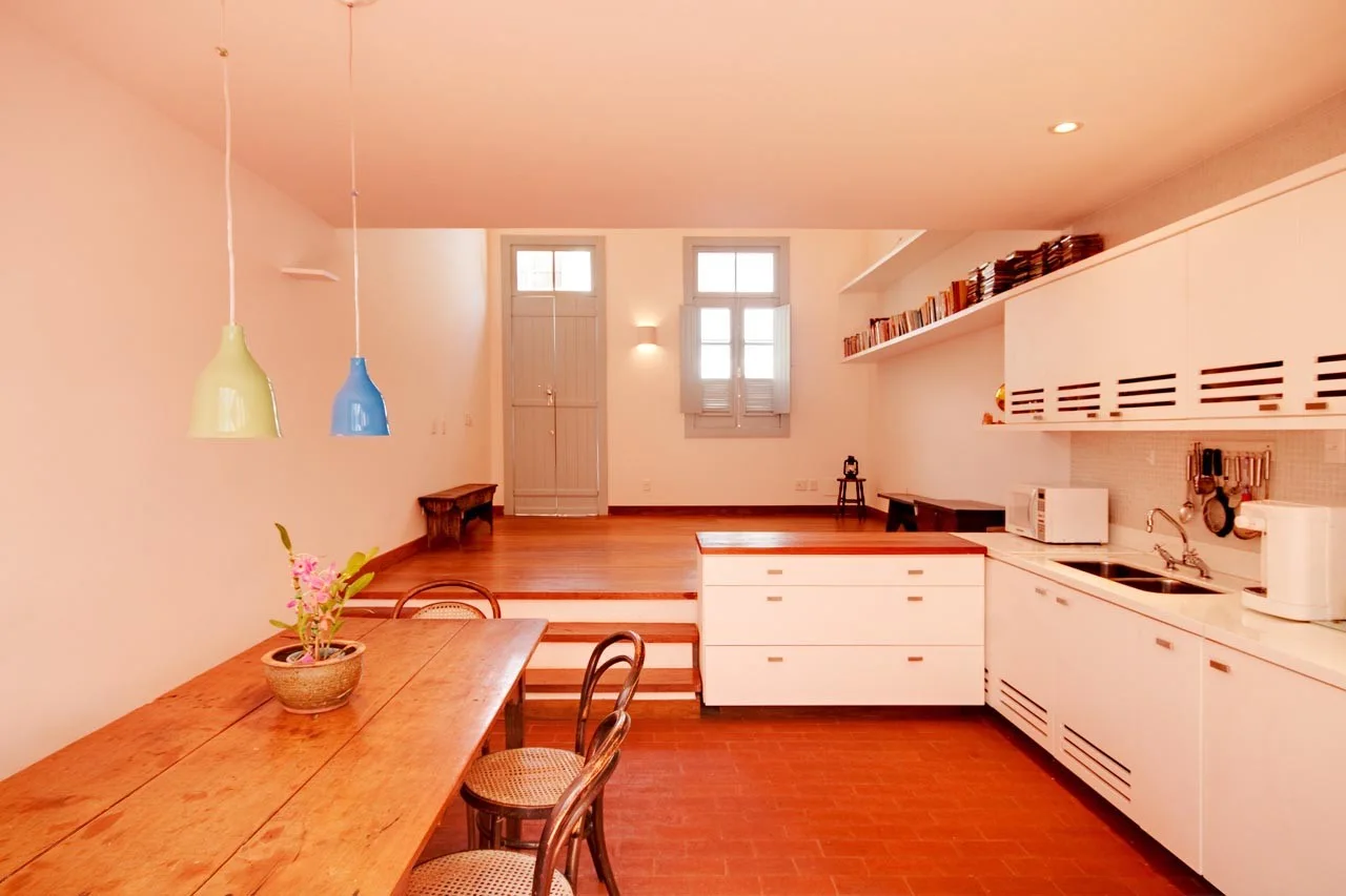 A split-level kitchen and dining room features a long wooden dining table, white minimalist cabinetry, terracotta tile floors, and colorful pendant lights.