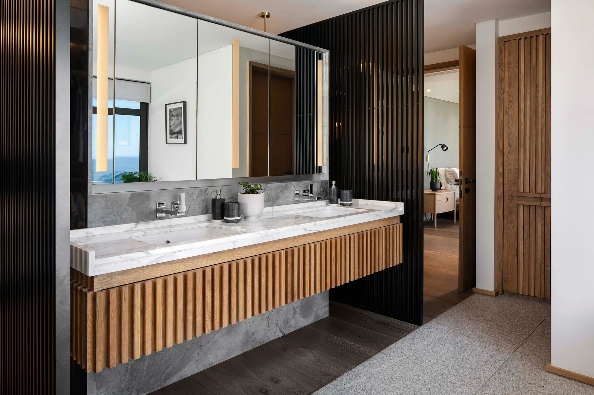 A modern double bathroom vanity features a wood-slatted cabinet base, a white marble countertop, and a large mirror flanked by integrated light strips.