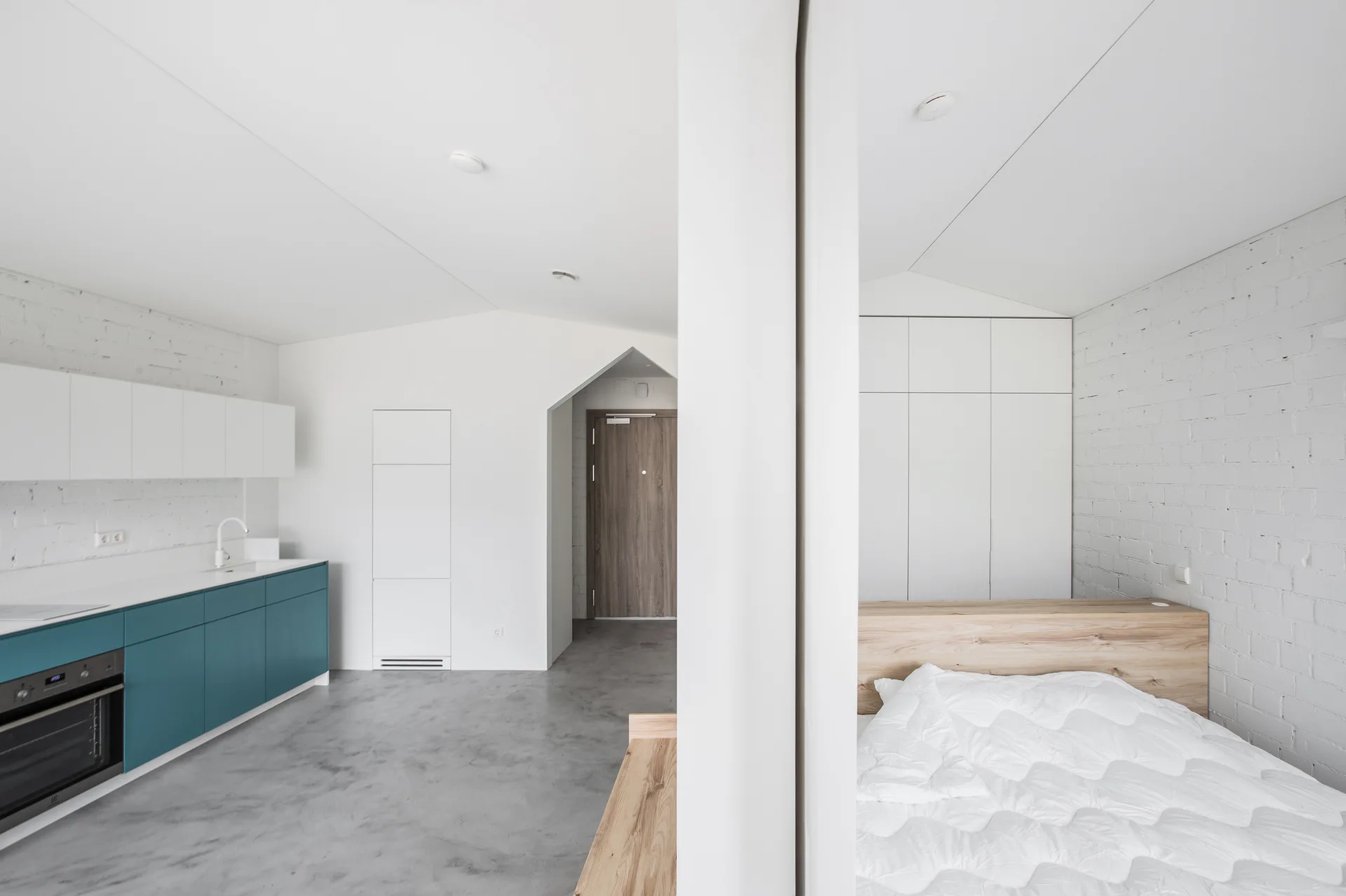 A minimalist studio apartment featuring a teal lower kitchen cabinet unit and a bed separated by a white vertical partition wall.
