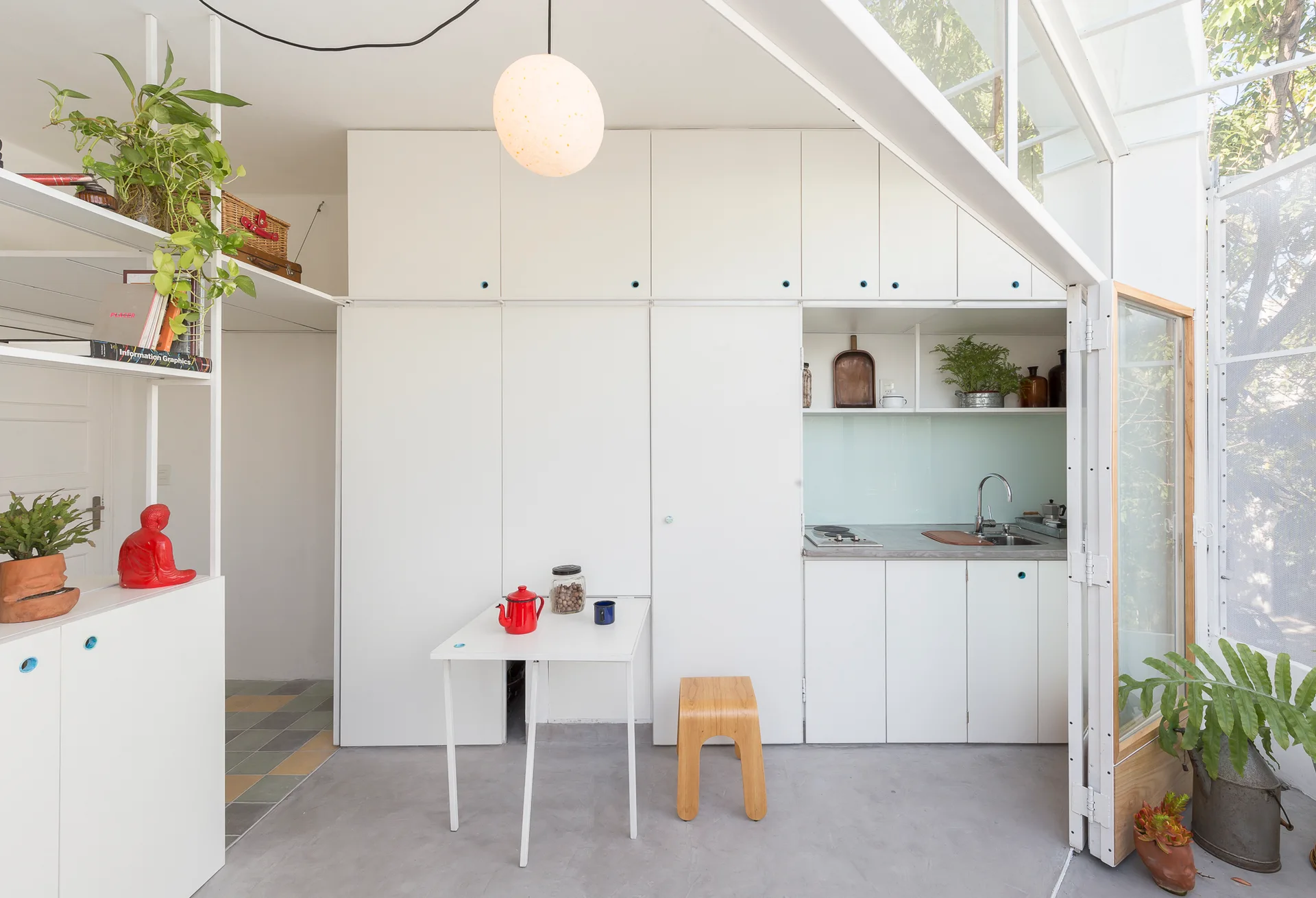 A minimalist white kitchenette featuring built-in wall cabinets, a small dining table, a wooden stool, and a smooth concrete floor.
