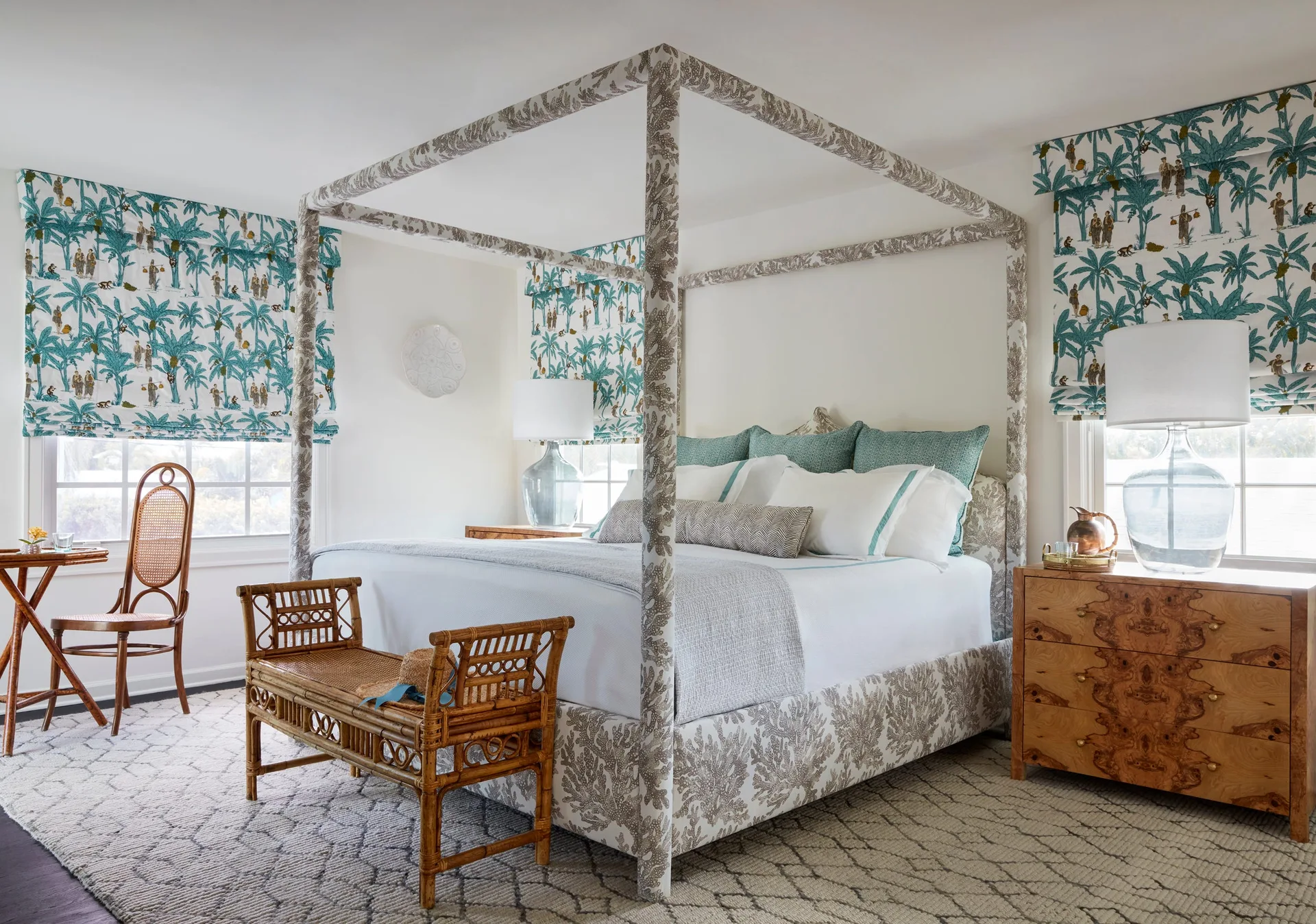 A bedroom features a four-poster canopy bed upholstered in coral-motif fabric, flanked by windows with teal patterned shades and a burl wood nightstand.