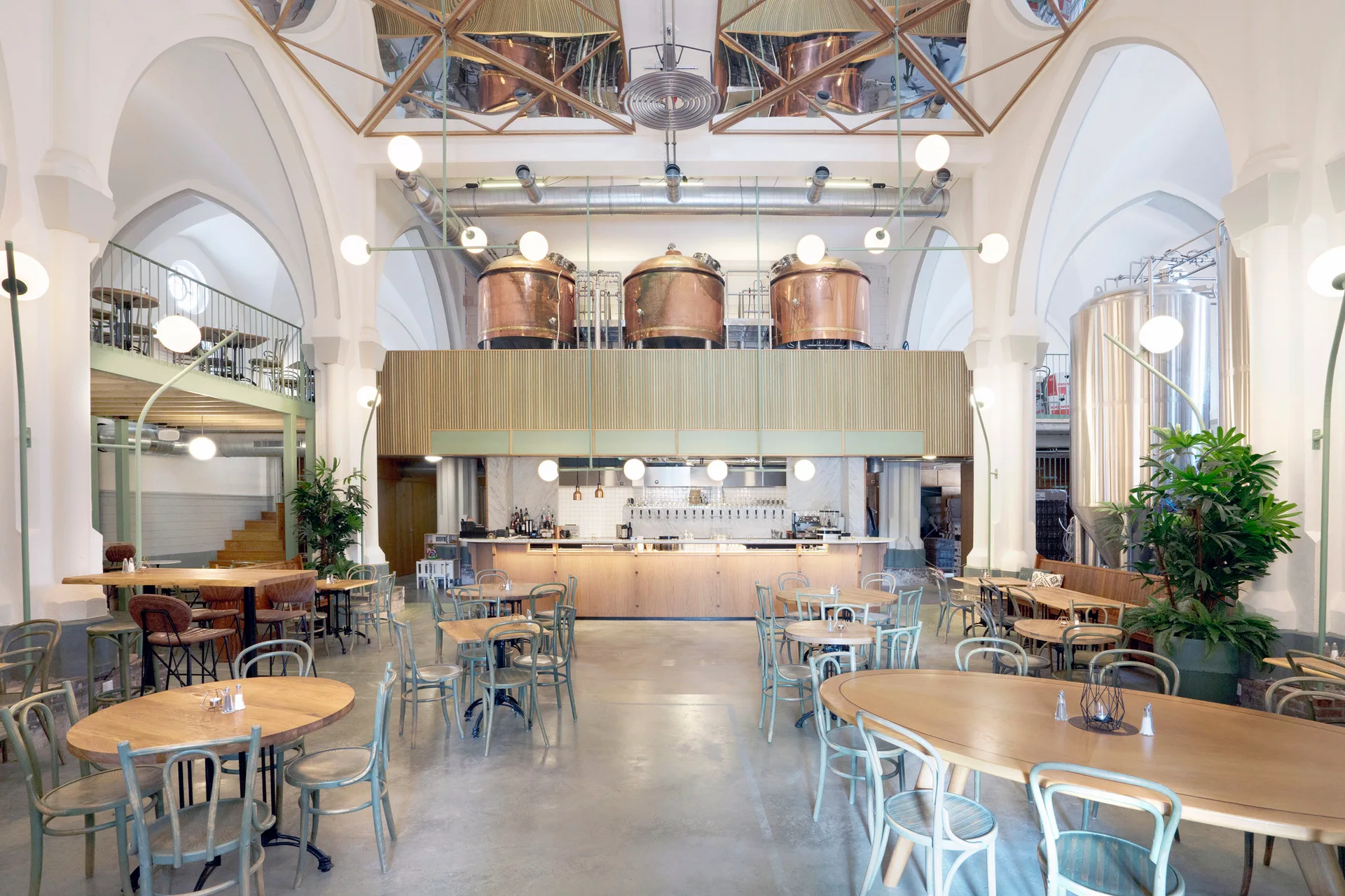 A spacious industrial brewery dining room features vaulted white ceilings, raised copper tanks, light wood tables, and a central wooden bar counter.