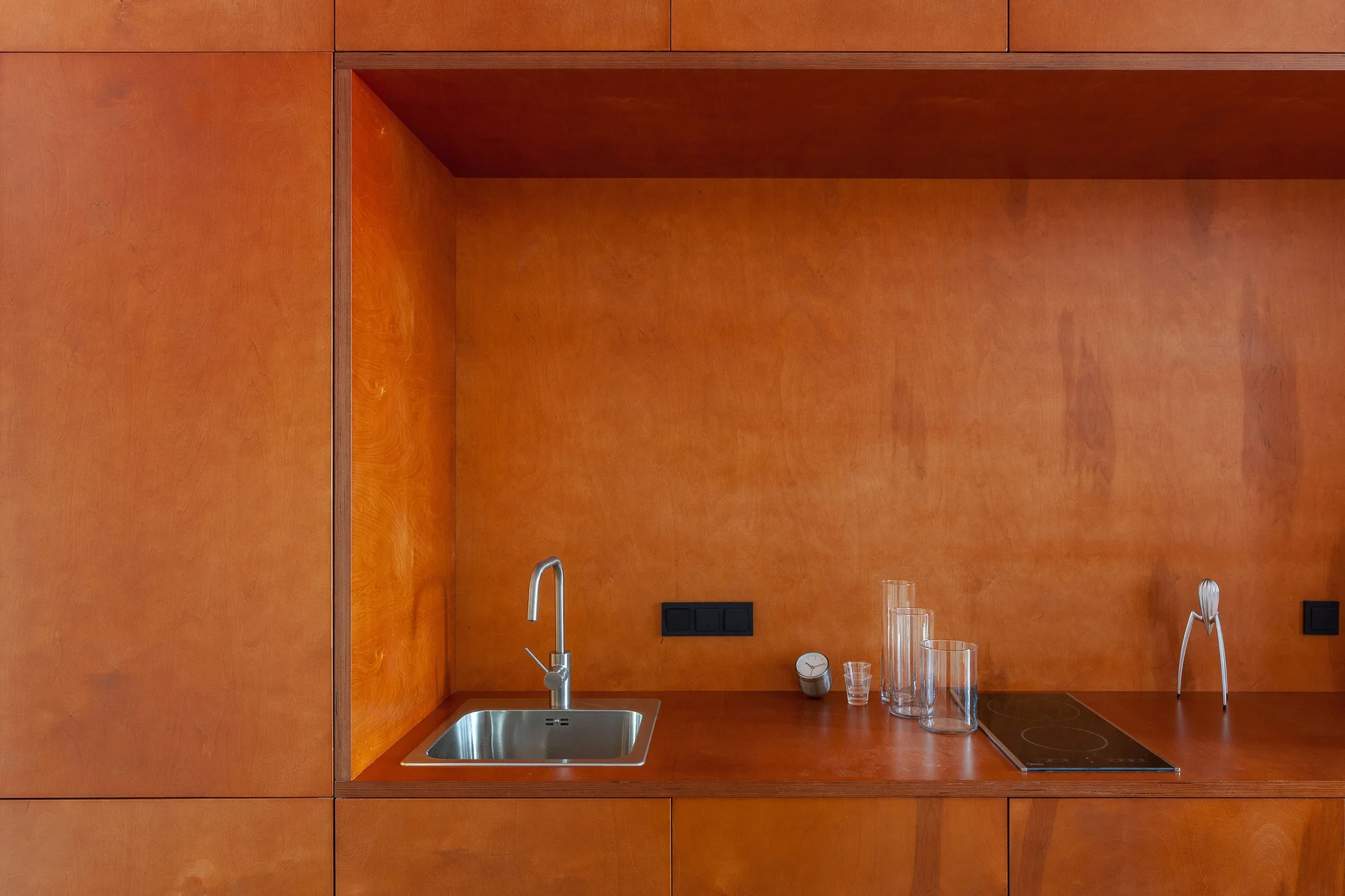 A built-in kitchen niche wrapped in warm orange-brown plywood features an integrated stainless steel sink and a black induction cooktop.