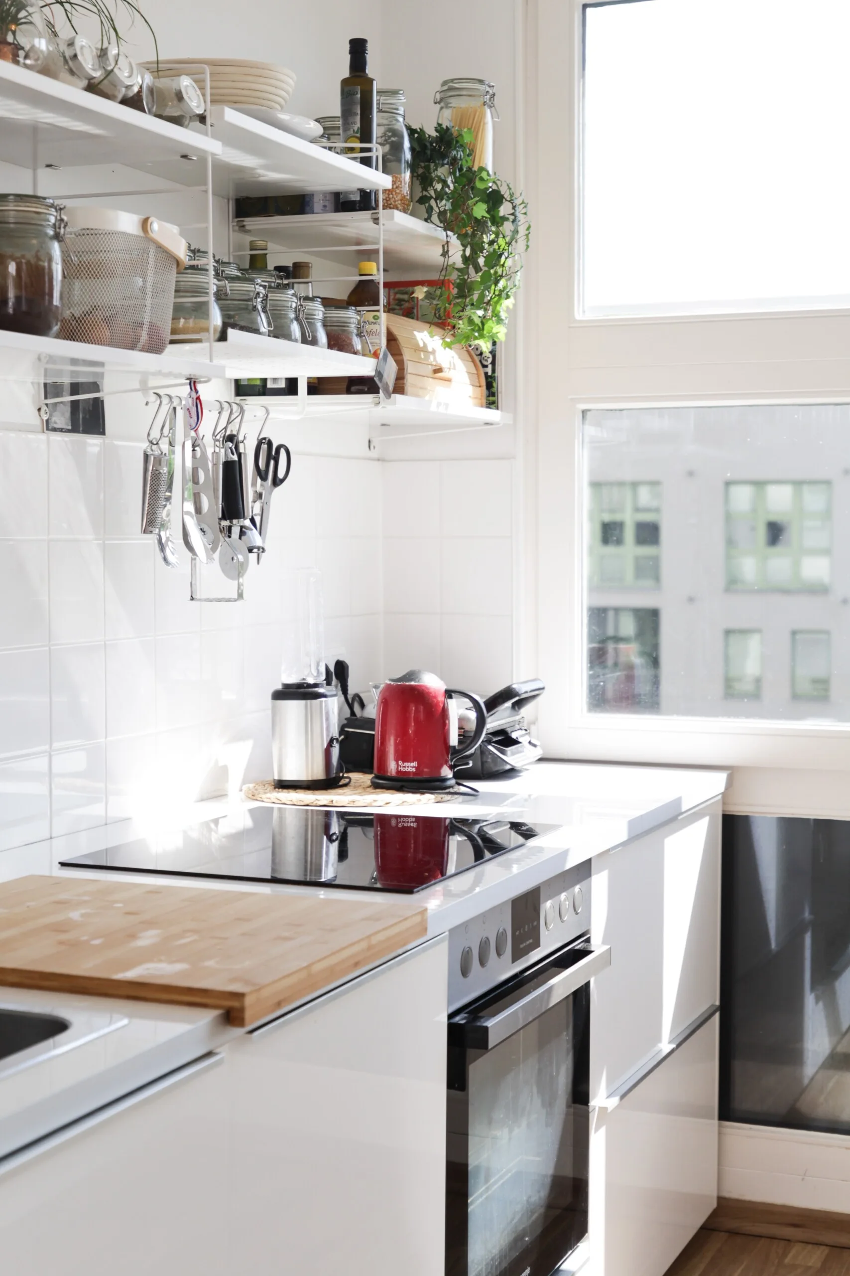 A bright white kitchen counter with a built-in oven, black induction cooktop, open wire shelving, and a red kettle next to a window.